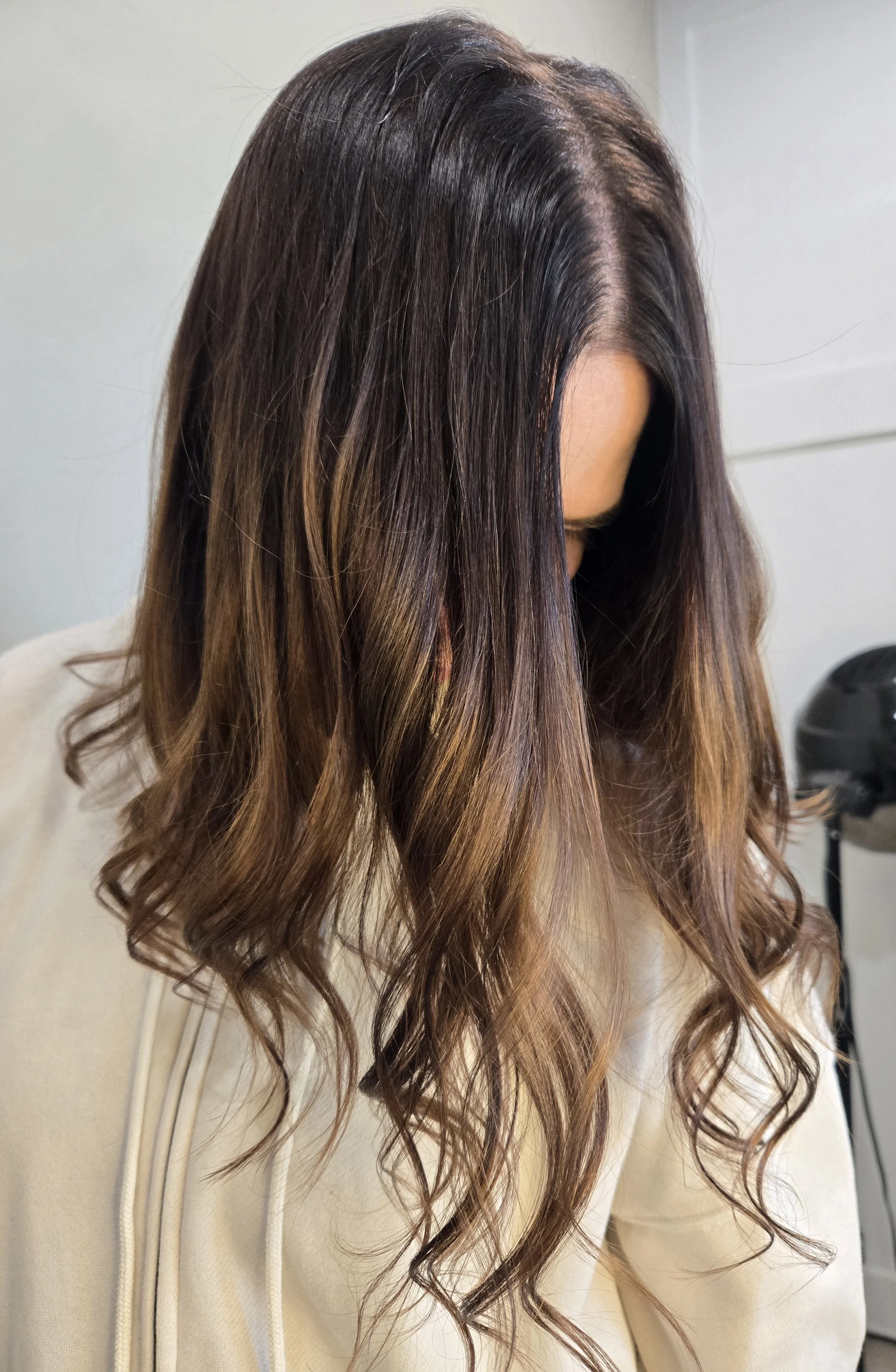 Photo of a woman with shoulder-length wavy hair colored in shades of brown, with darker roots fading into lighter tips, looking downward in a salon or hair studio.