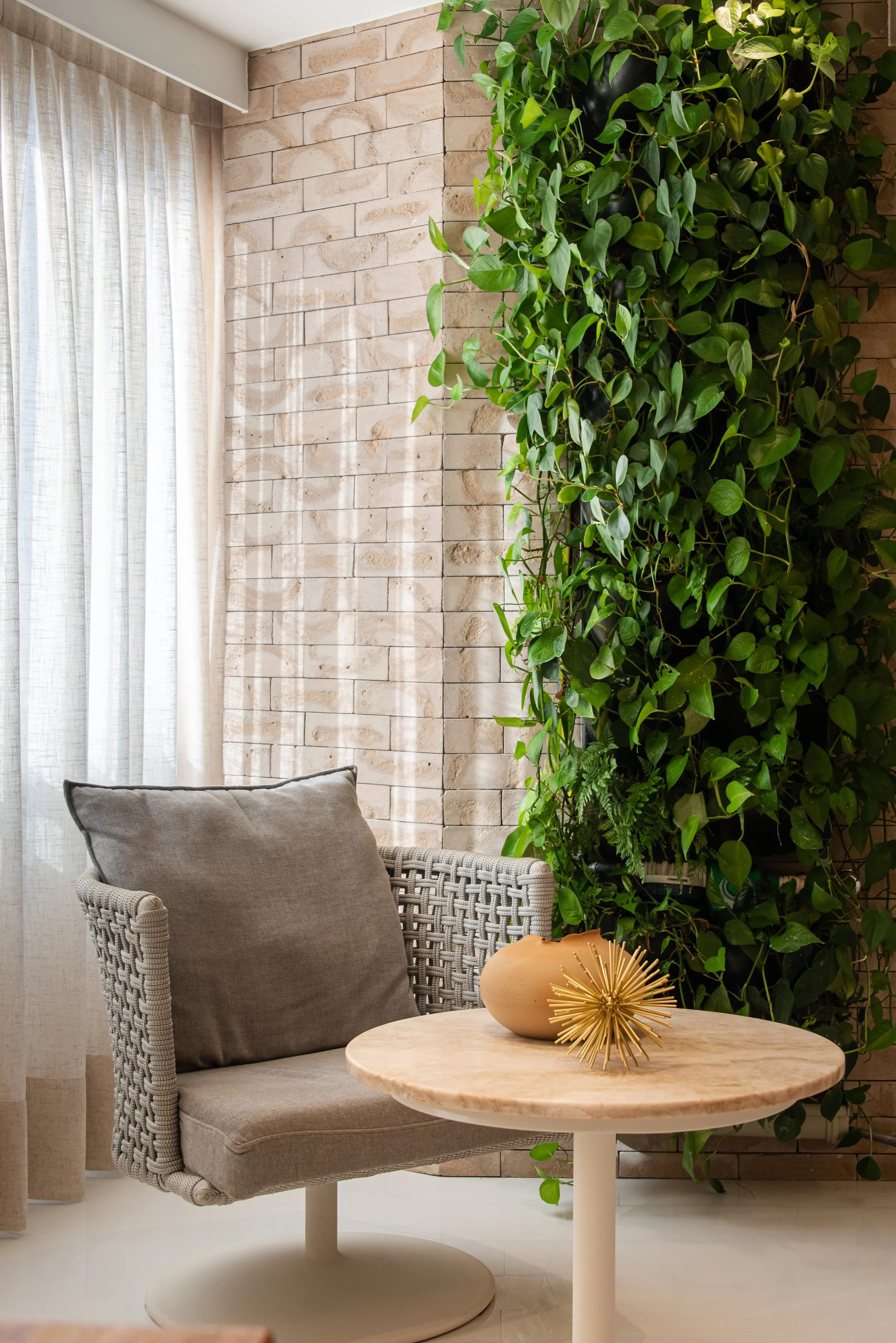 A cozy corner with a woven armchair with a gray cushion, a round marble-top table with decorative objects, green hanging plants, a brick wall, and sheer white curtains.