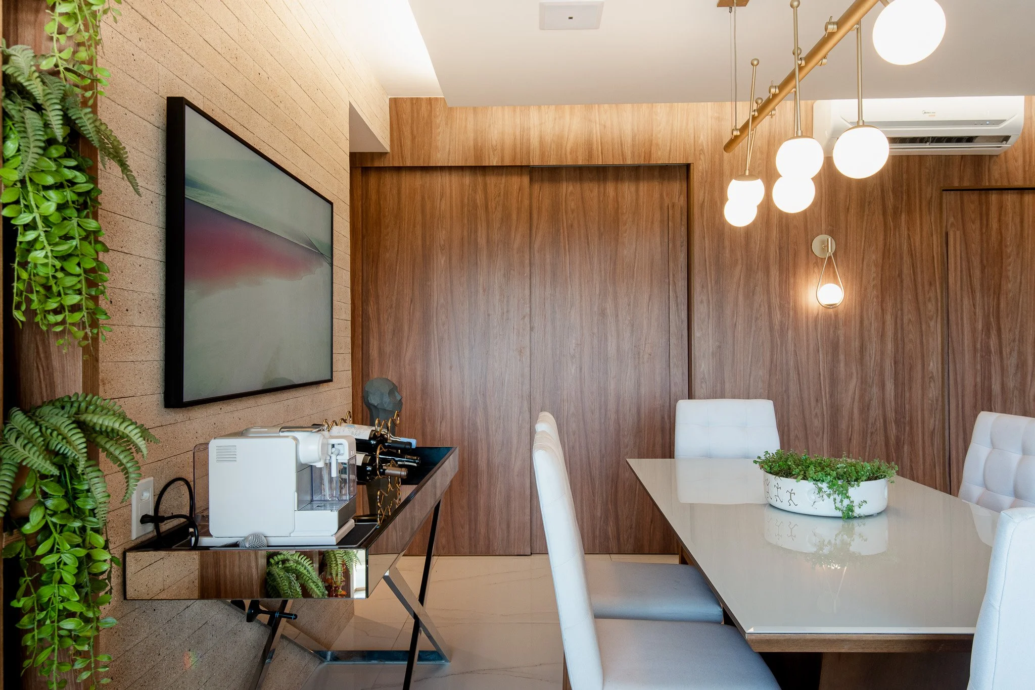 Modern dining room with a wooden wall, white chairs at a glossy table, and pendant lights, with a small side table holding a beverage station and a green plant in a white pot.