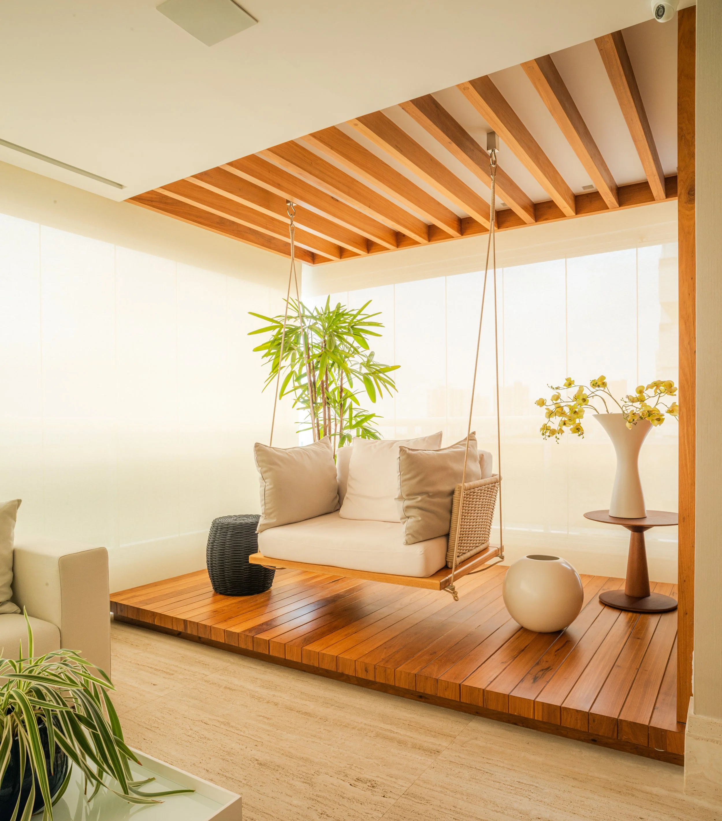 A modern indoor space with a wooden platform featuring a hanging sofa with beige cushions, a large potted plant, a white decorative vase on a side table, a round white vessel, and sheer window coverings with natural light.