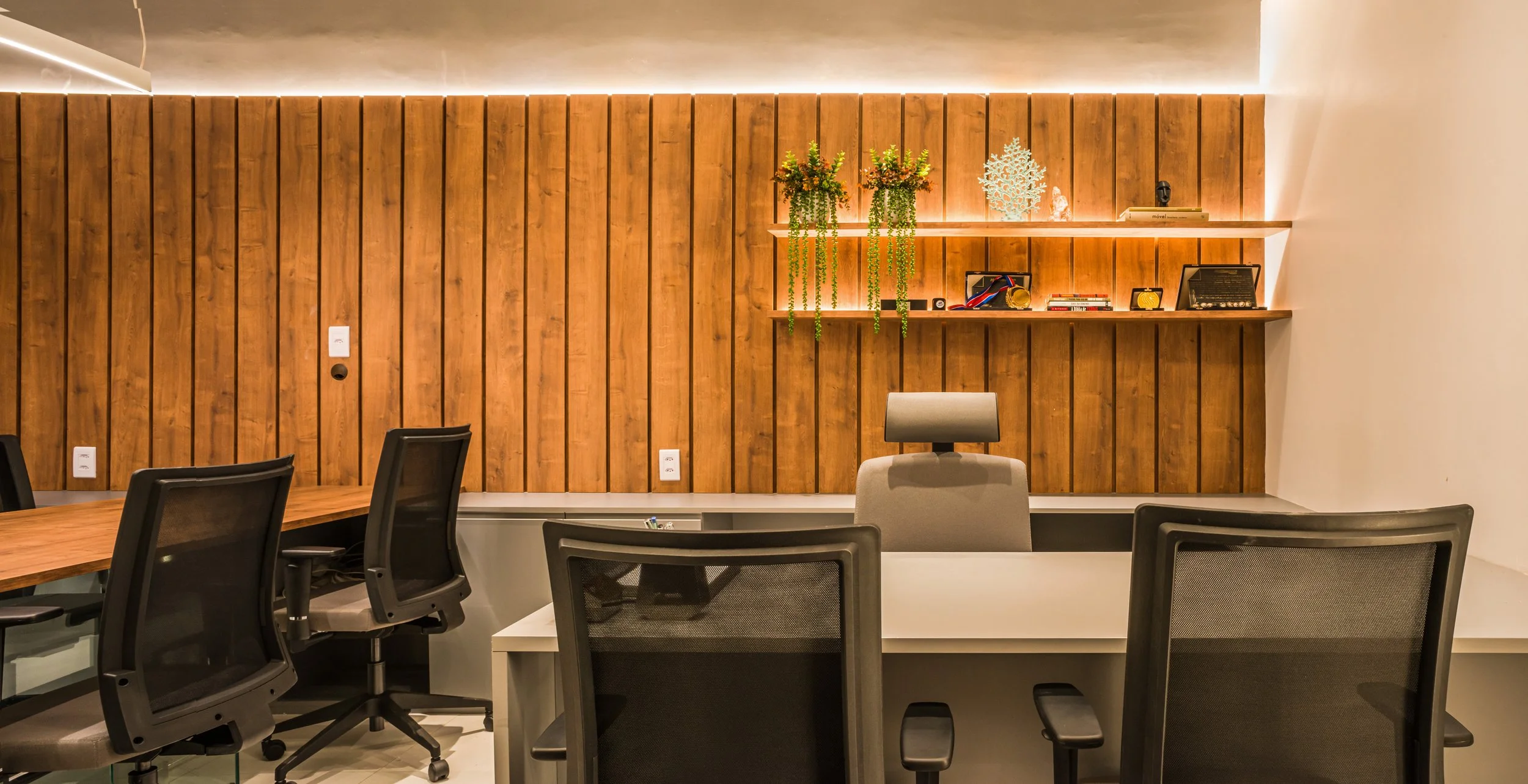 Modern office space with a wooden accent wall, office chairs, and a desk. Shelves on the wall hold decorative plants, awards, books, and decorative items, illuminated by warm lighting.