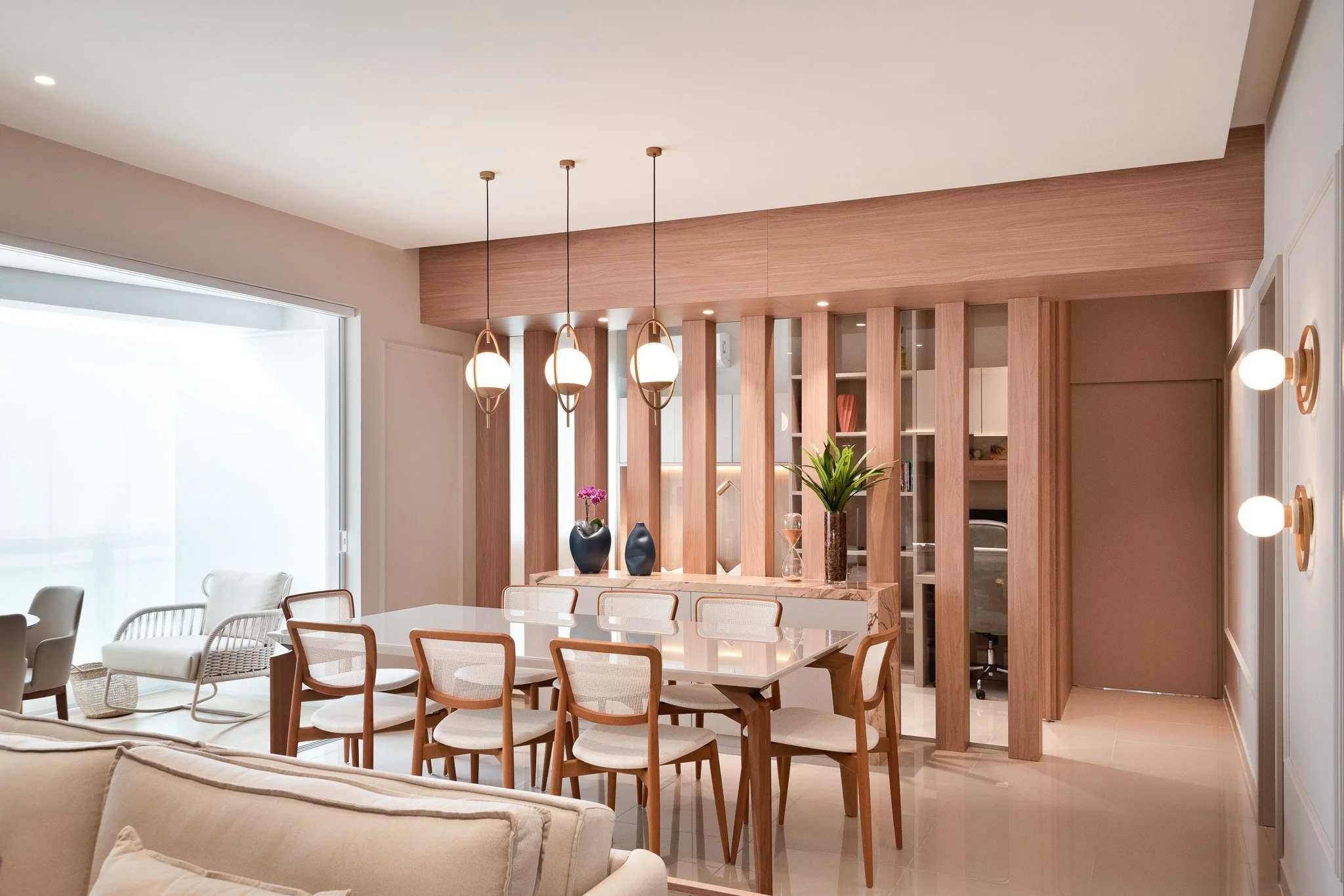 Modern dining room with a marble table, wooden chairs, hanging pendant lights, decorative vases with flowers and plants, wooden slat partition, and a bright window.