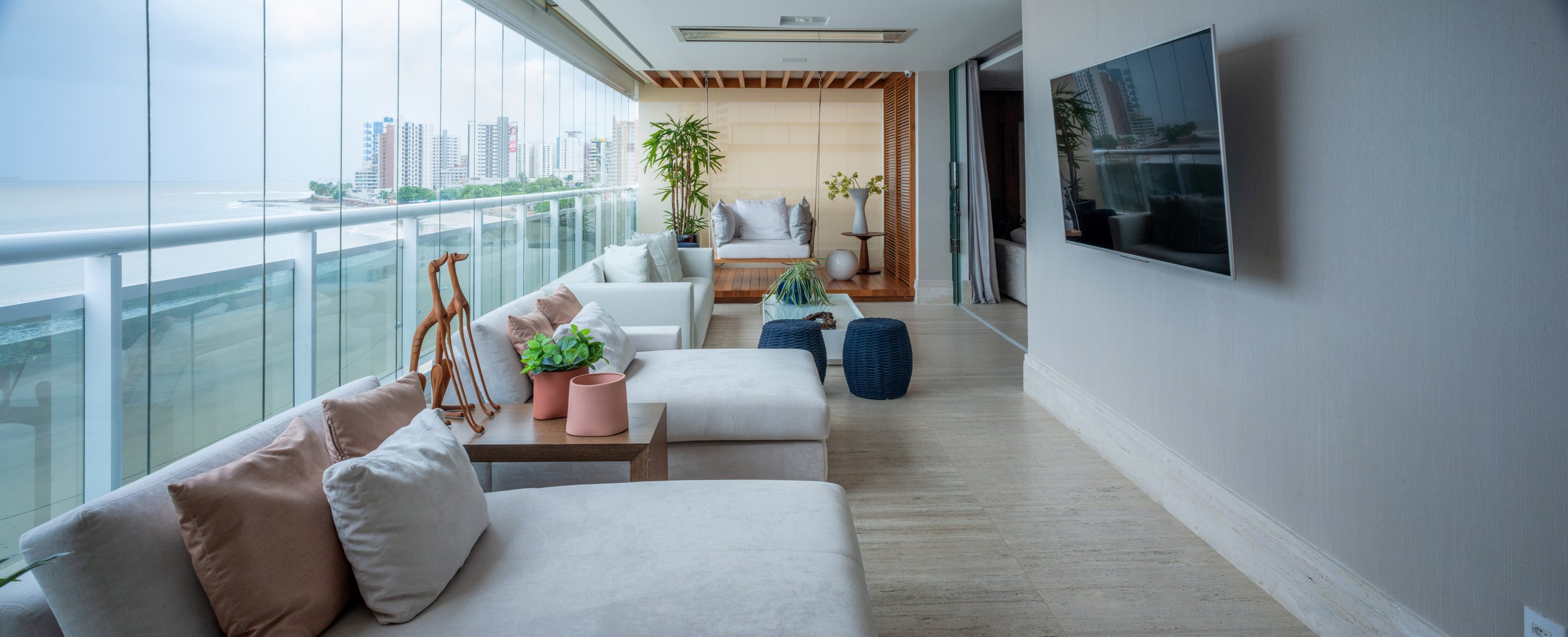 Modern living room with large glass windows overlooking city buildings, featuring white sofas, potted plants, a wall-mounted TV, and a wooden covered seating area at the end.