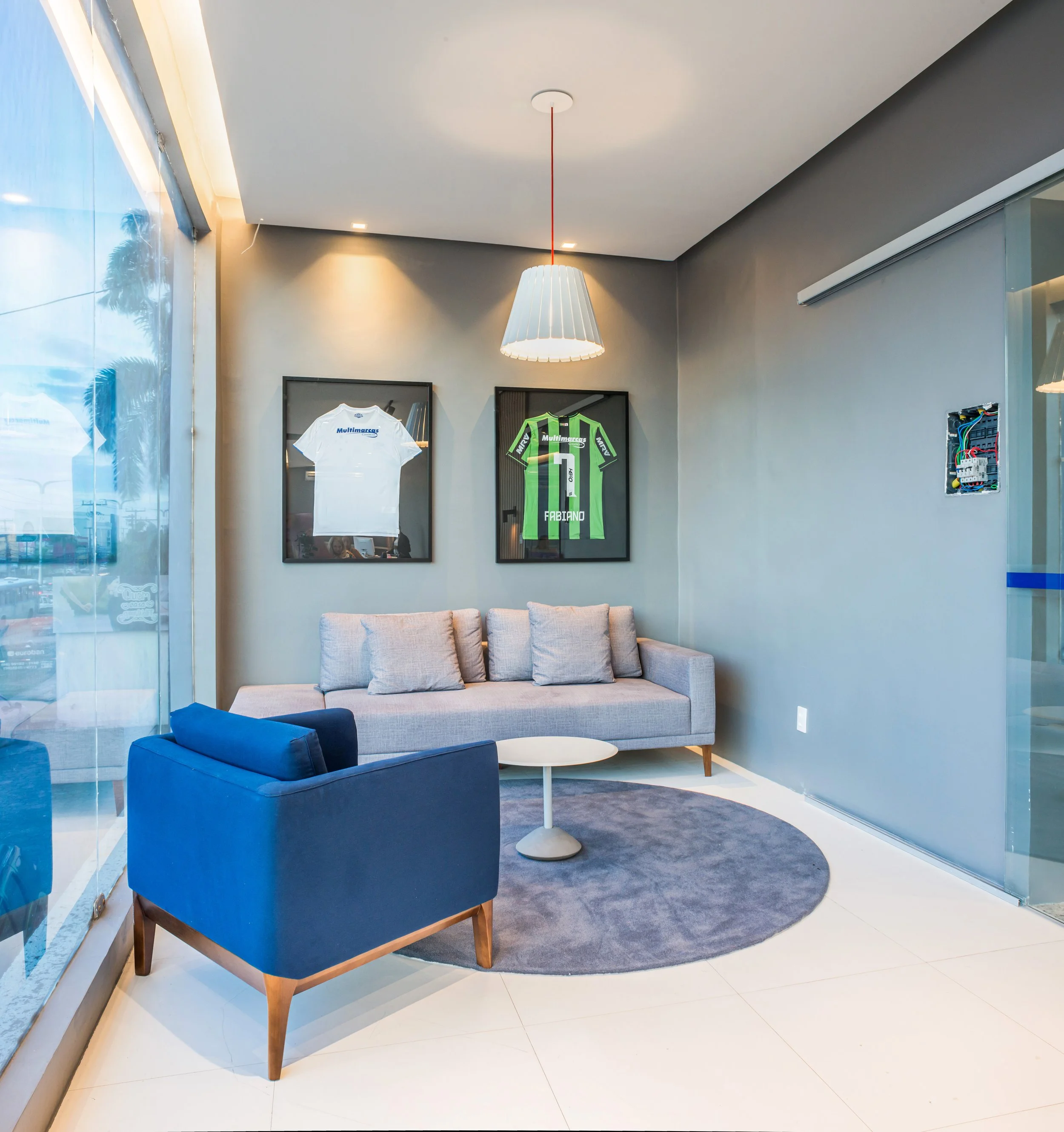 Modern lounge area with a grey sofa, a blue armchair, and a white round coffee table on a circular grey rug. Two framed sports jerseys, one white and one green, are hung on the wall behind the sofa. A large window with a view of palm trees and a blue