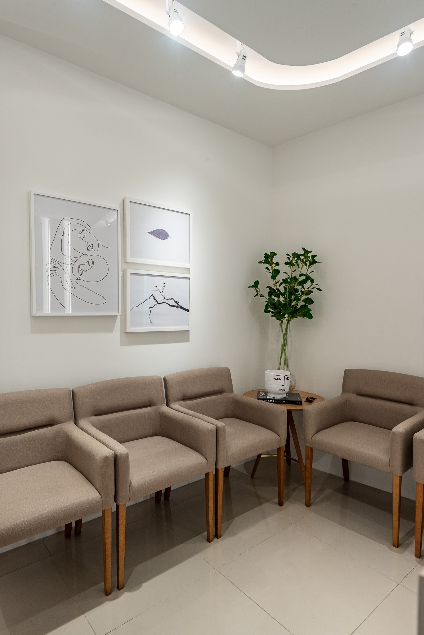 A waiting room with four beige chairs and a small wooden side table holding a white decorative face-shaped vase and a stack of books, next to a tall green plant. The white walls are decorated with three minimalist art prints and the ceiling has white