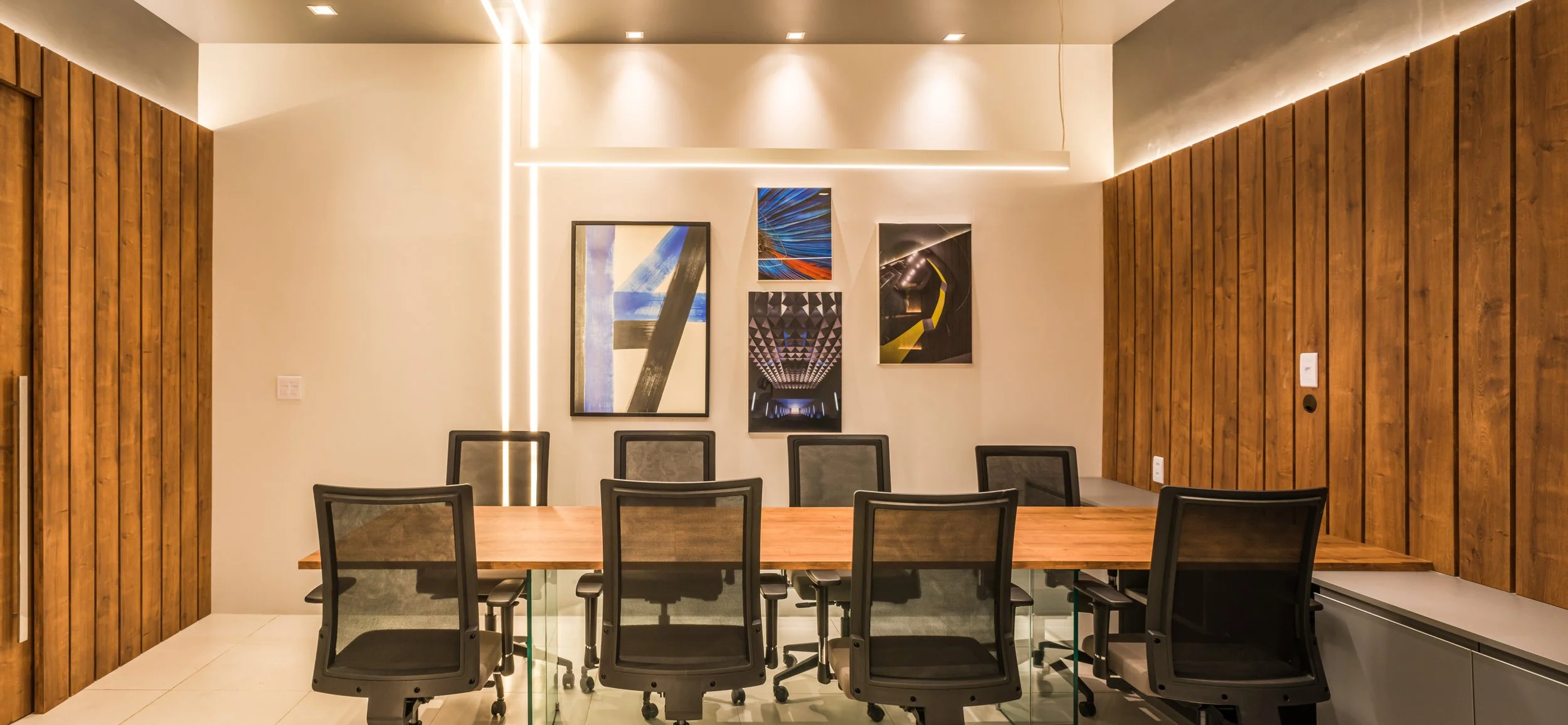 Modern conference room with a long wooden table, eight black office chairs, and artwork on the wall featuring city and architectural images. The walls are wood-paneled with subtle lighting.