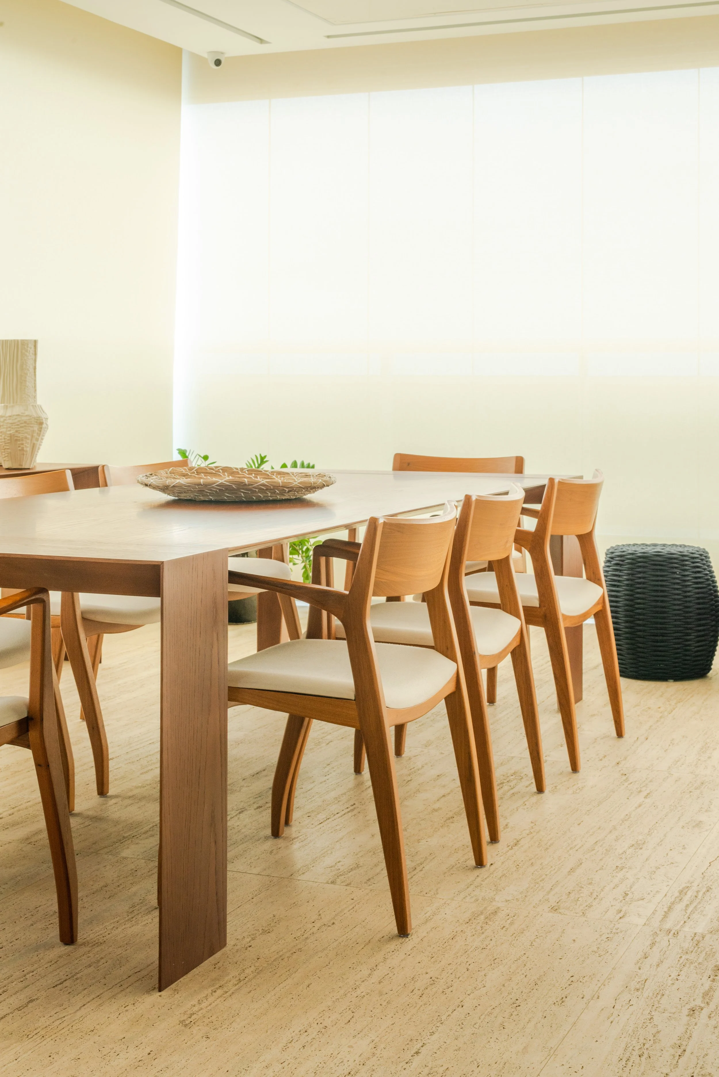 A modern dining room with wooden chairs around a large wooden table, decorated with a woven centerpiece. The room has bright, natural lighting from large windows with light-colored blinds. There is a black woven pouf on the right and a potted plant b