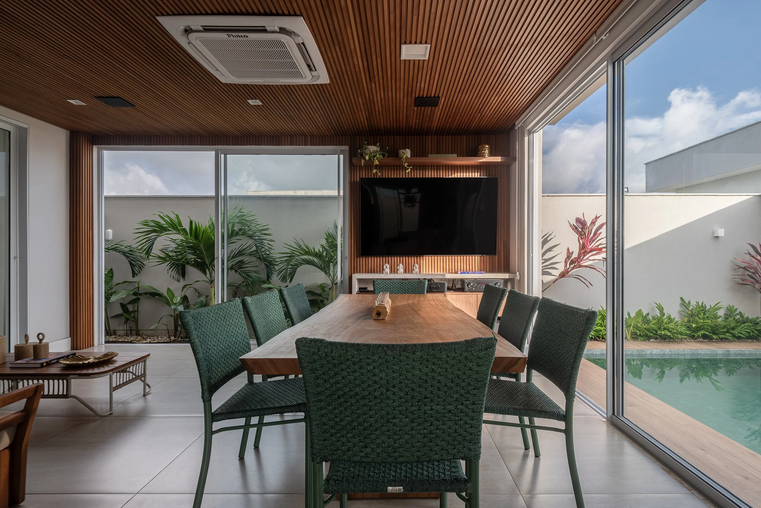 Modern dining room with a wooden table, green chairs, indoor plants, a wall-mounted TV, and large sliding glass doors leading to a patio with a pool and outdoor plants.