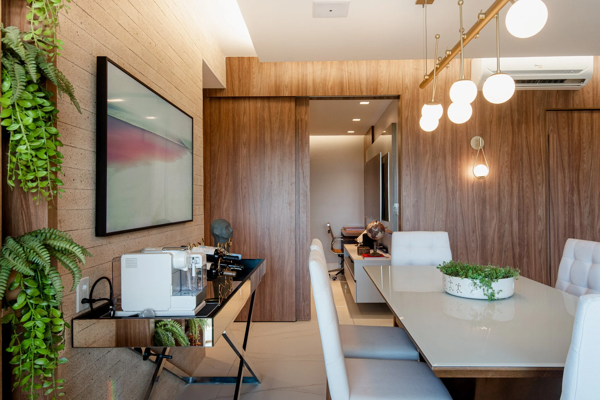 Modern dining room with wooden walls, white chairs, a glass-top table with a plant centerpiece, hanging globe lights, and a coffee machine on a black console table.