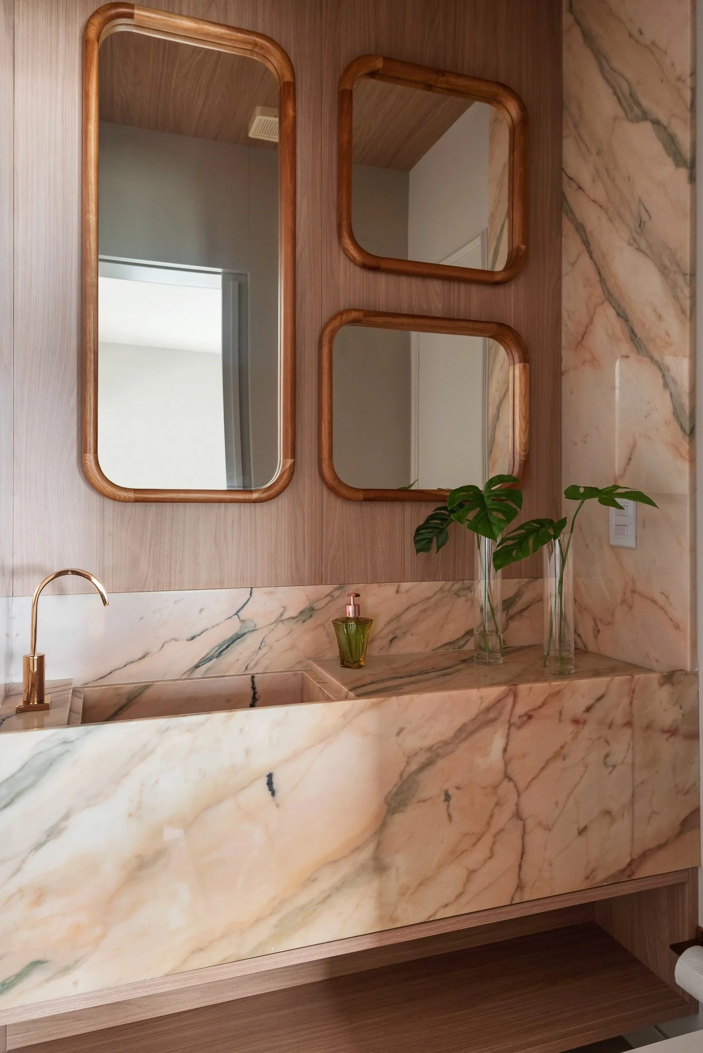 Bathroom with marble countertops and backsplash, wooden-framed mirrors, a gold faucet, a small green soap dispenser, and vases with green leaves on the countertop.
