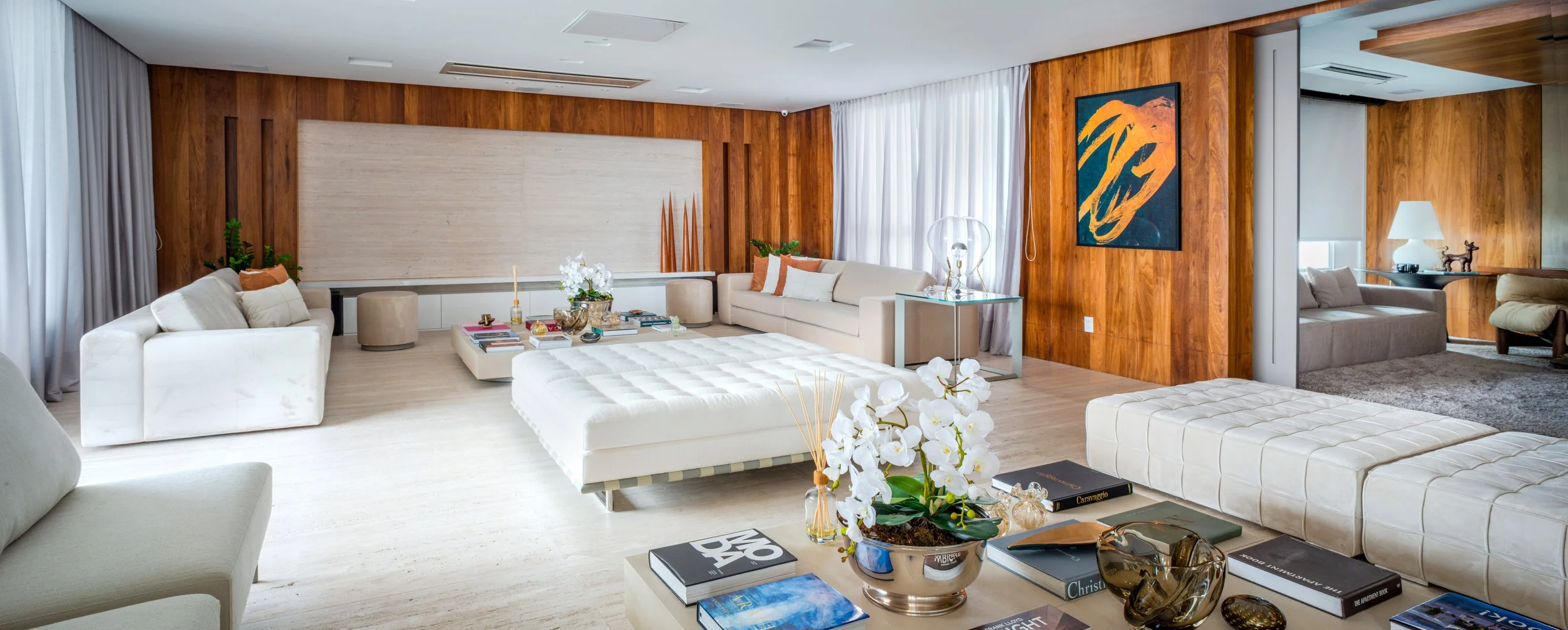 Luxurious living room with cream and beige sofas, wooden wall paneling, and modern art on the wall. Decor includes potted plants, artwork, books, a glass table lamp, and floral arrangements.