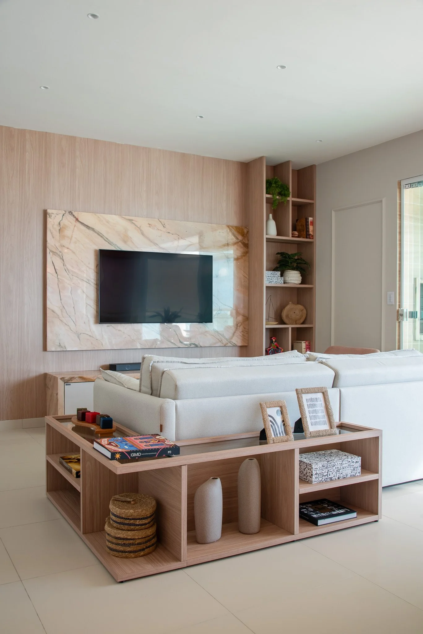 Modern living room with a wall-mounted TV, white sofas, and a wooden shelf with decorative items and books.