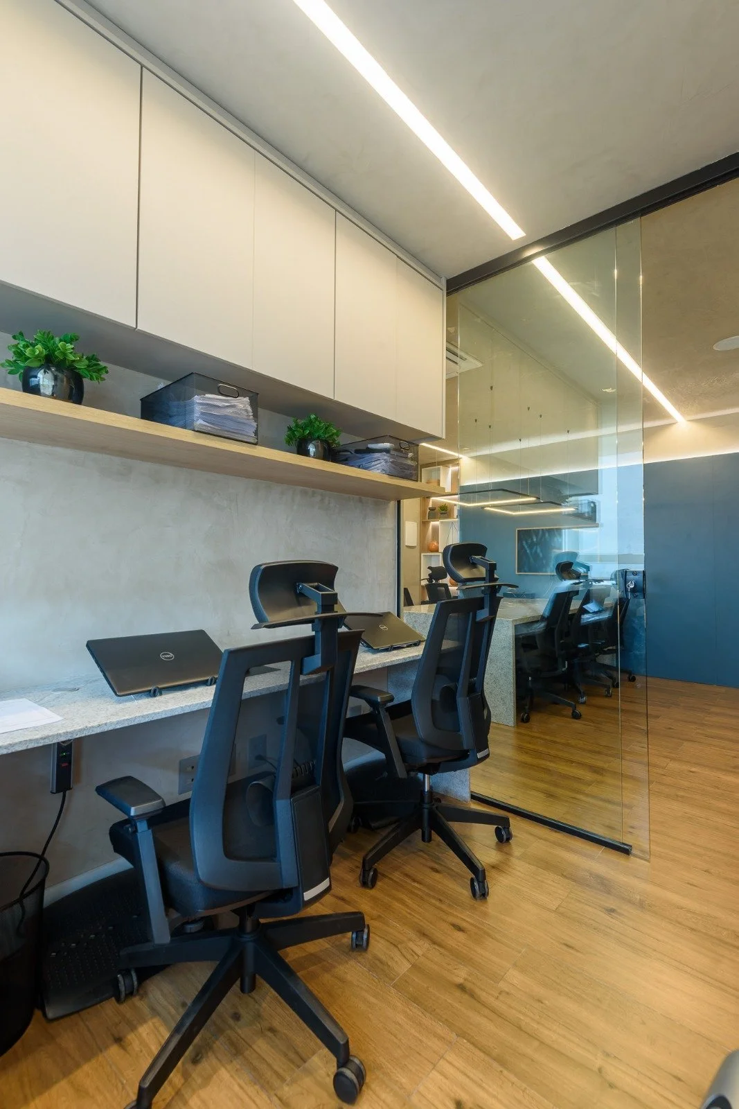 Modern office workspace with four black ergonomic chairs, laptops, a glass wall, and wooden flooring.