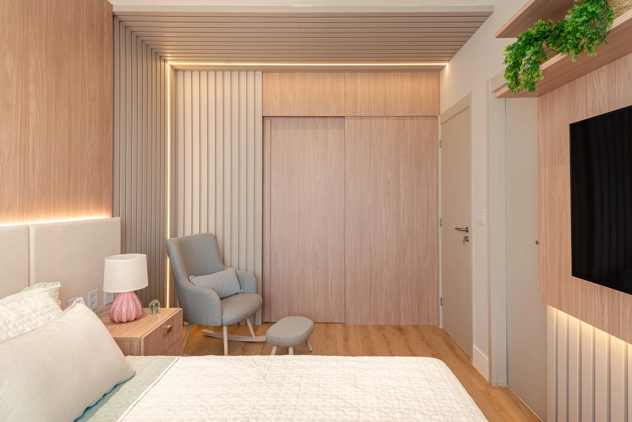 A modern bedroom with light wood paneling on the walls and ceiling, a bed with white bedding, a nightstand with a pink lamp, a gray armchair with a matching ottoman, and a wall-mounted flat-screen TV. The room also features vertical lighting accents 