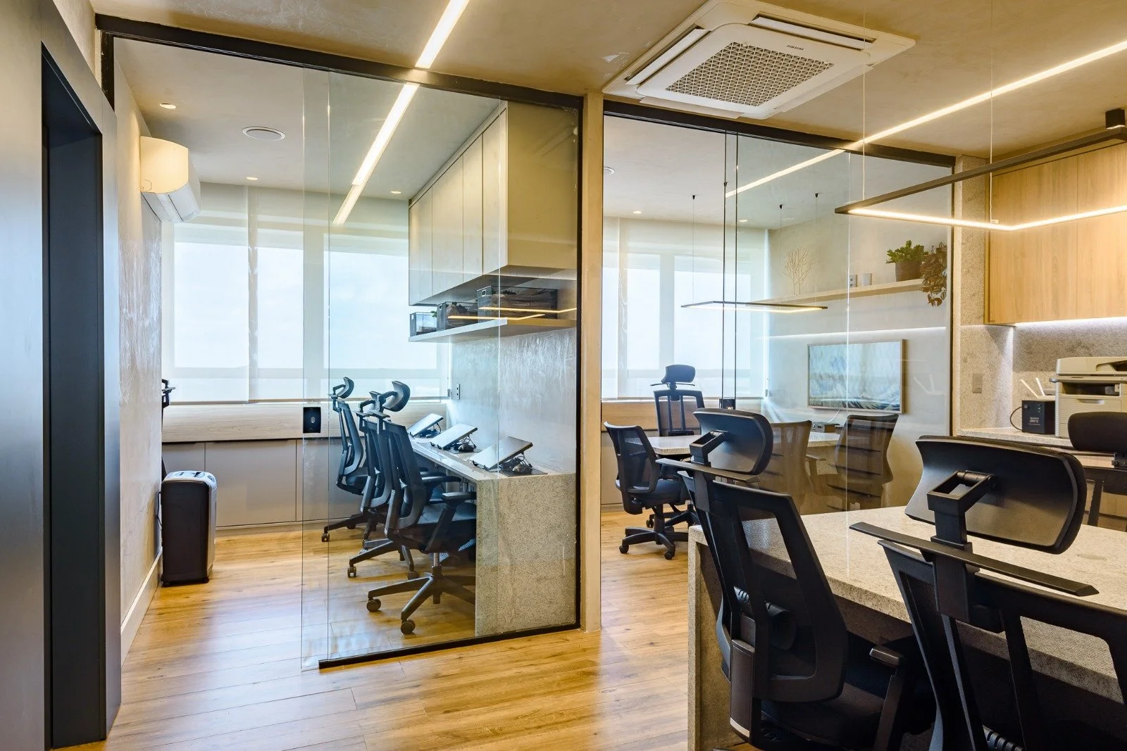 Modern office space divided by glass walls, featuring desks with ergonomic chairs, large windows with blinds, wooden flooring, and ceiling lighting.