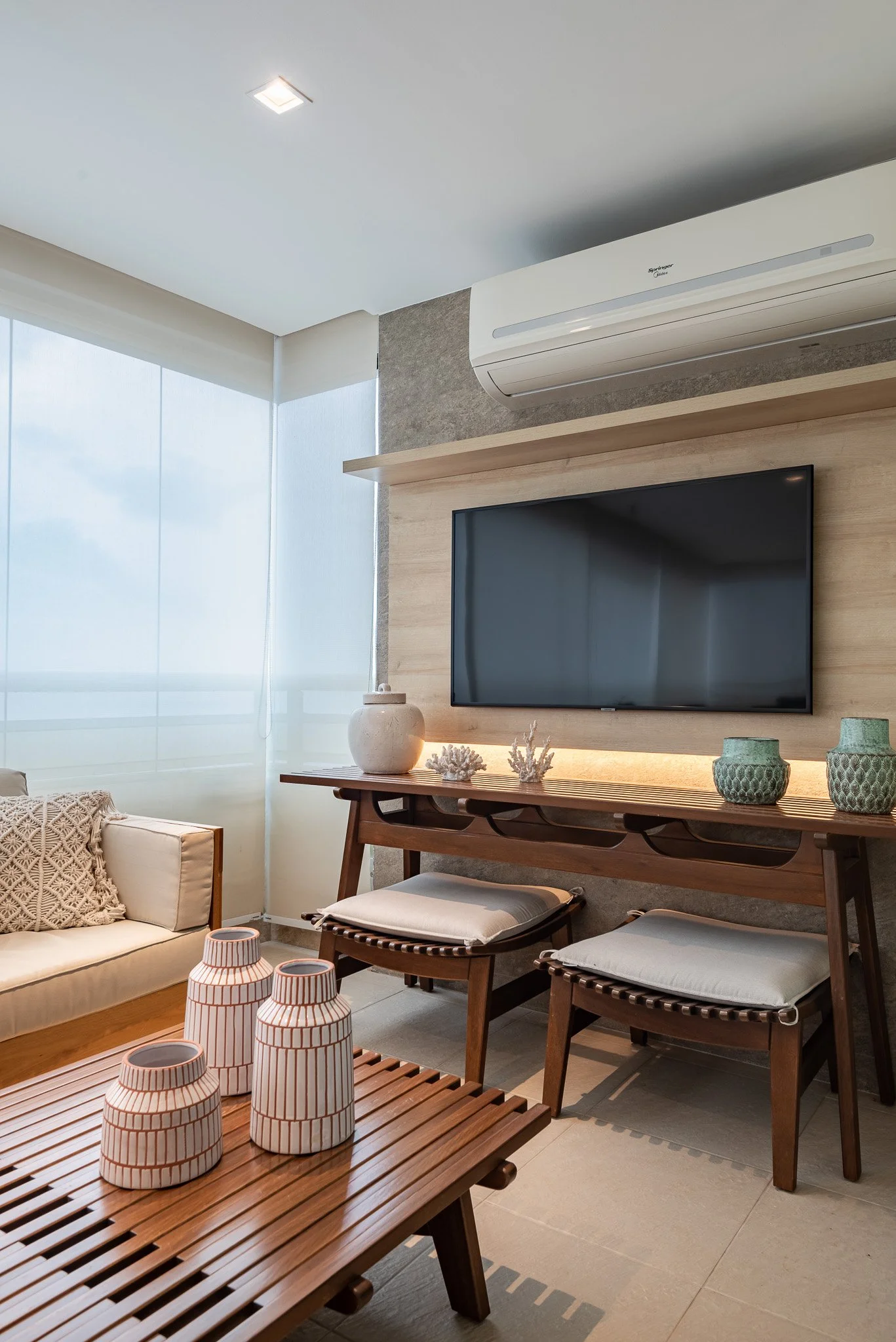 Living room with a wall-mounted TV, air conditioner, wooden console table with decorative vases and coral, beige sofa with throw pillow, wooden coffee table with striped vases, large window with sheer curtains, and beige tiled flooring.