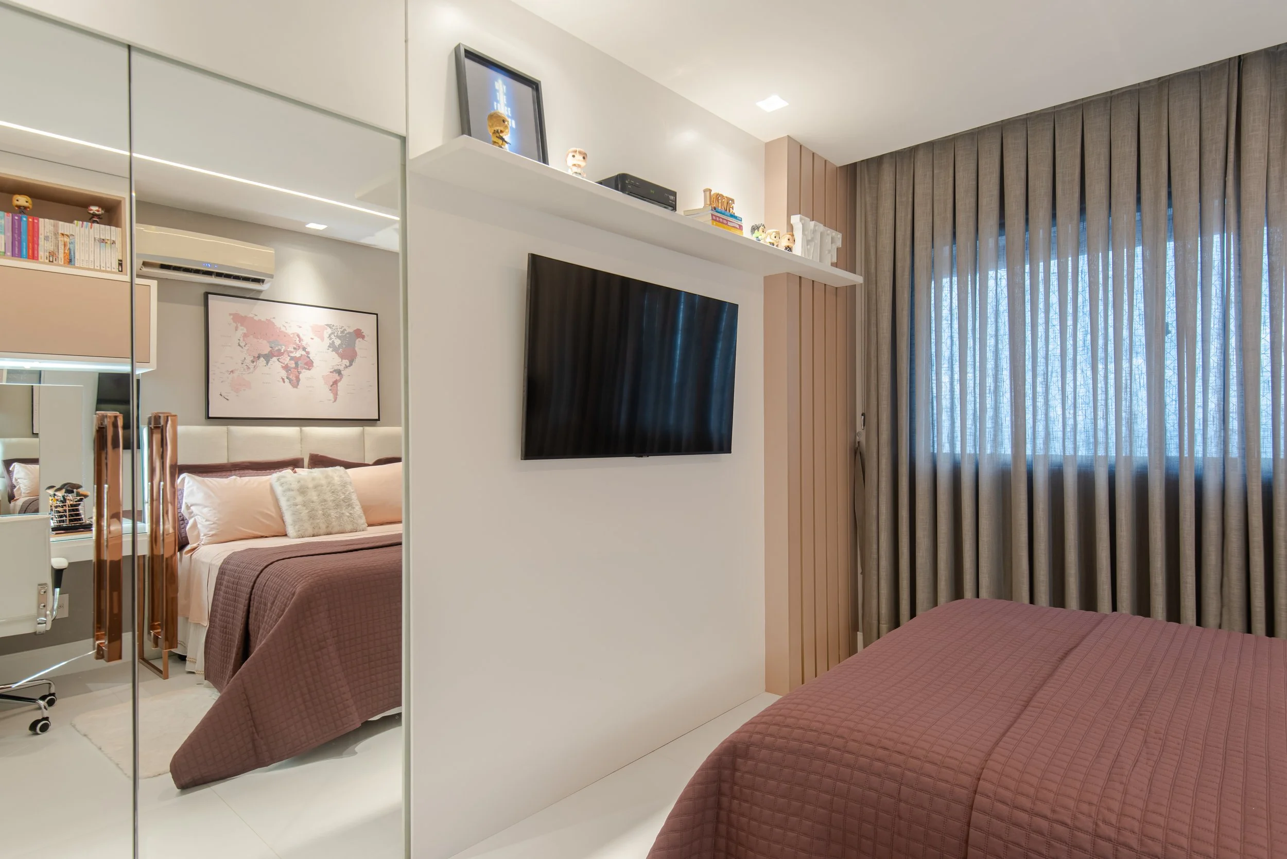 A modern bedroom with a bed, quilted bedspread, and pillows, reflected in mirrored closet doors. Another bedroom with a bed against a wall is visible through an open doorway, with a world map on the wall. A wall-mounted TV is on a partition wall, wit