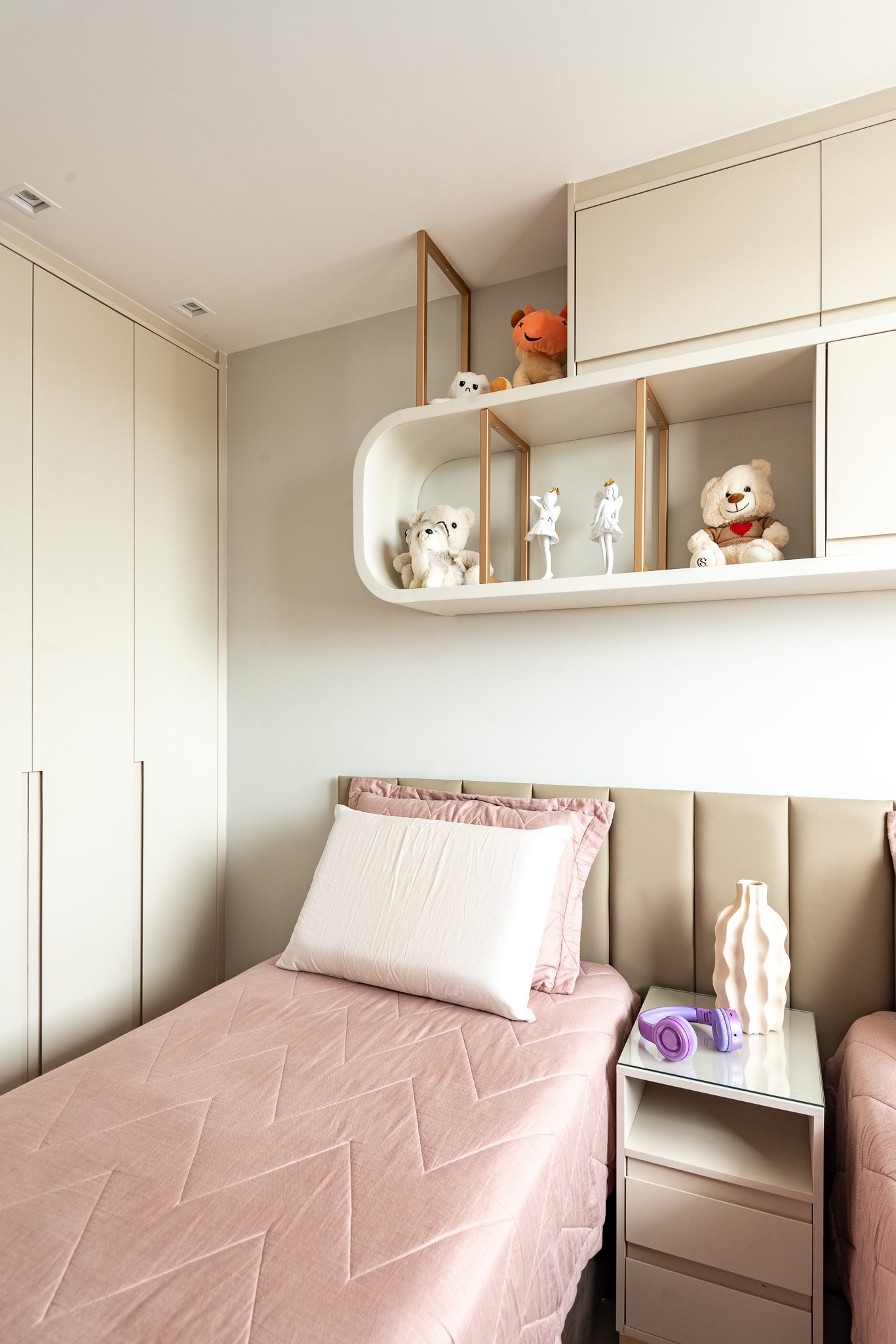 A neatly made bed with a pink quilt and white pillows in a girls' bedroom. A white nightstand with purple headphones and a cream-colored vase is beside the bed. Above, a shelf with teddy bears, plush toys, and ballet figurines decorates the wall.