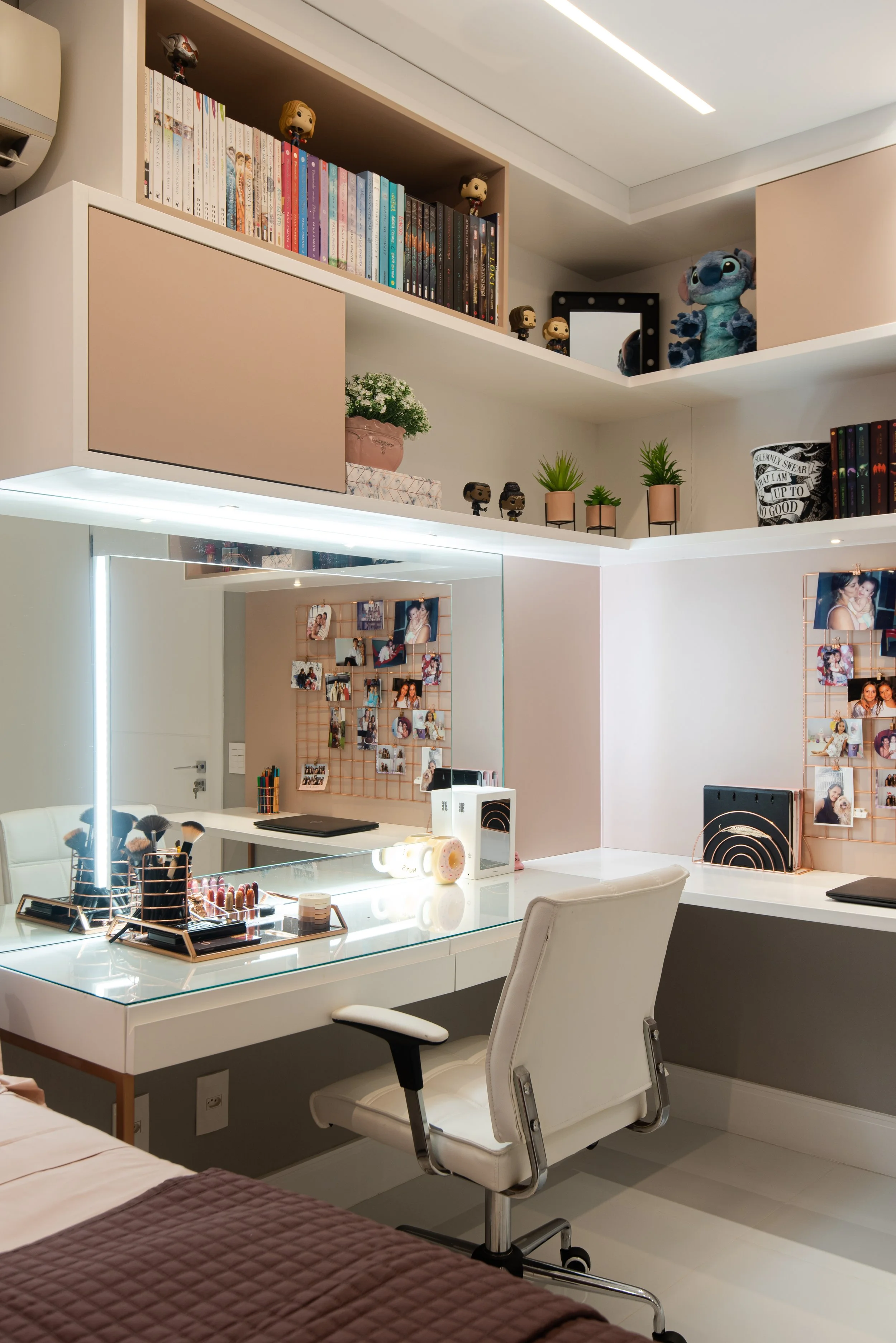 A modern bedroom corner with a white desk, a white office chair, a large mirror with LED lighting, and a wall with photo and item decorations. Books, figurines, plants, and fluffy toys are on shelves above the desk.
