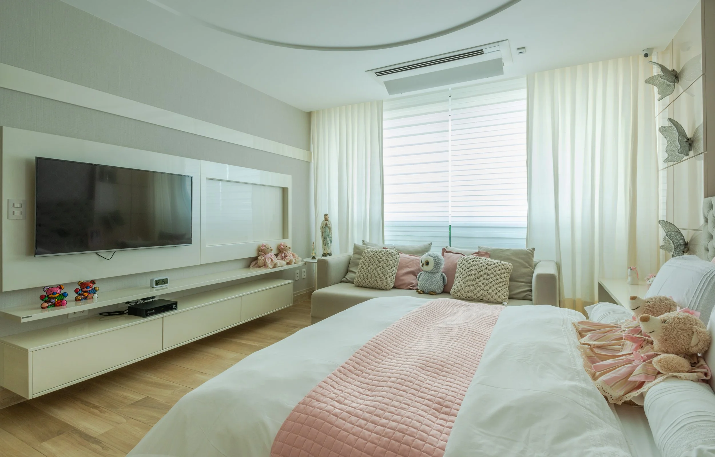 A bright, modern bedroom with a large bed, pillows, and teddy bears, a flat-screen TV on the wall, and a sofa with plush toys by the window covered in sheer curtains.