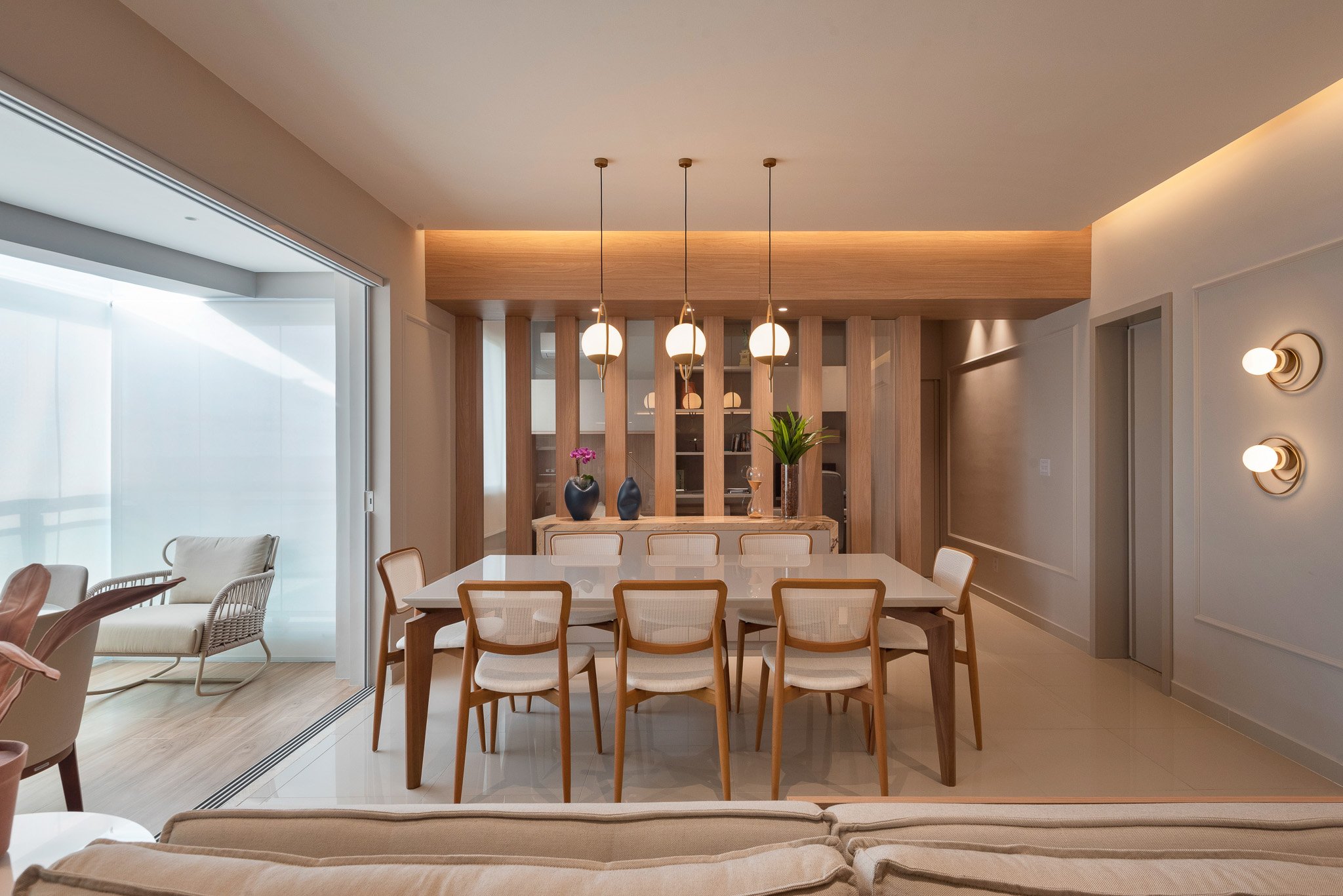 Modern dining room with a white table, eight beige chairs, wooden panels, pendant lights, decorative vases, and a comfortable sitting area with armchairs.