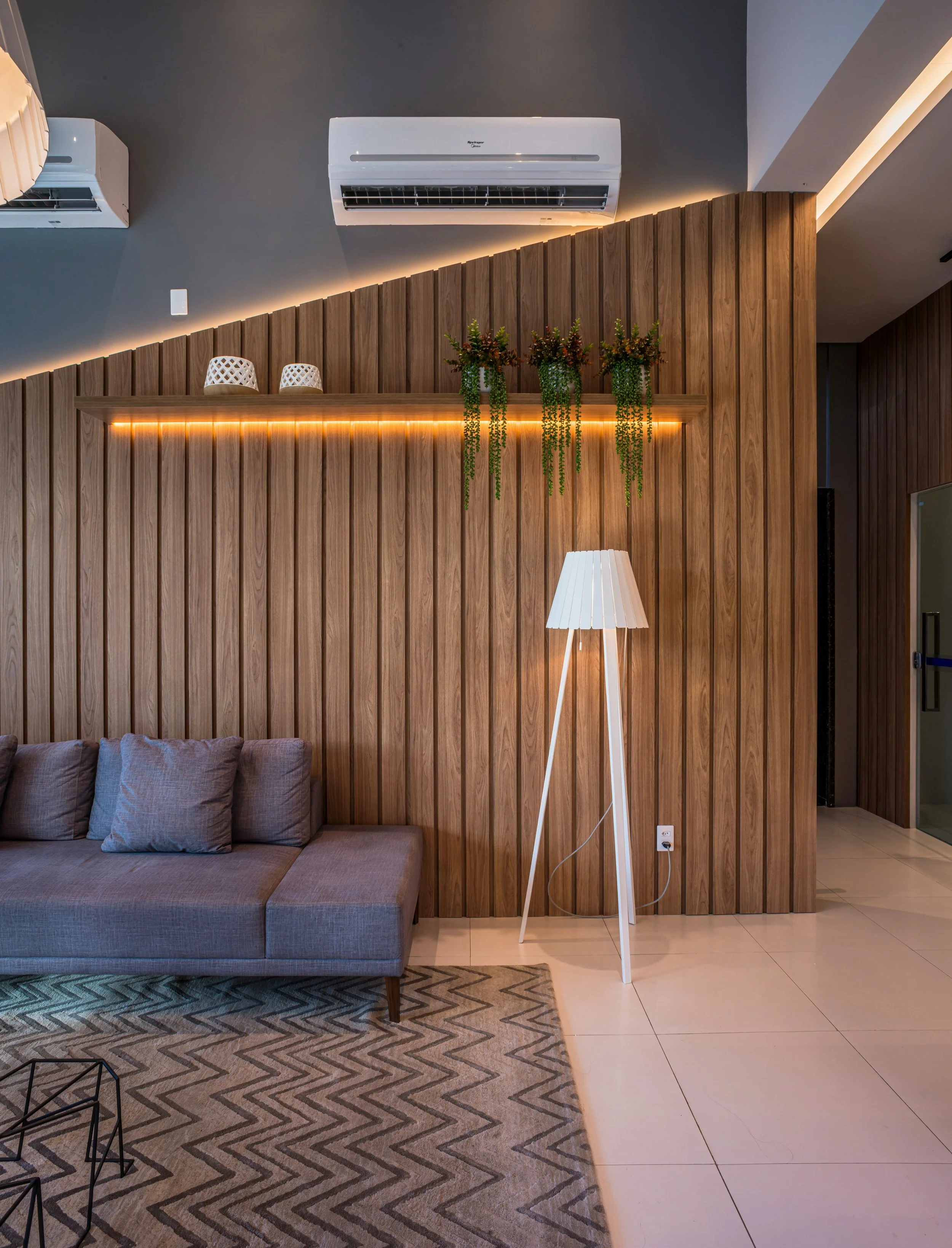 Modern interior living room with gray sofa, wooden wall paneling, a tall tripod floor lamp, and decorative plants on a shelf.