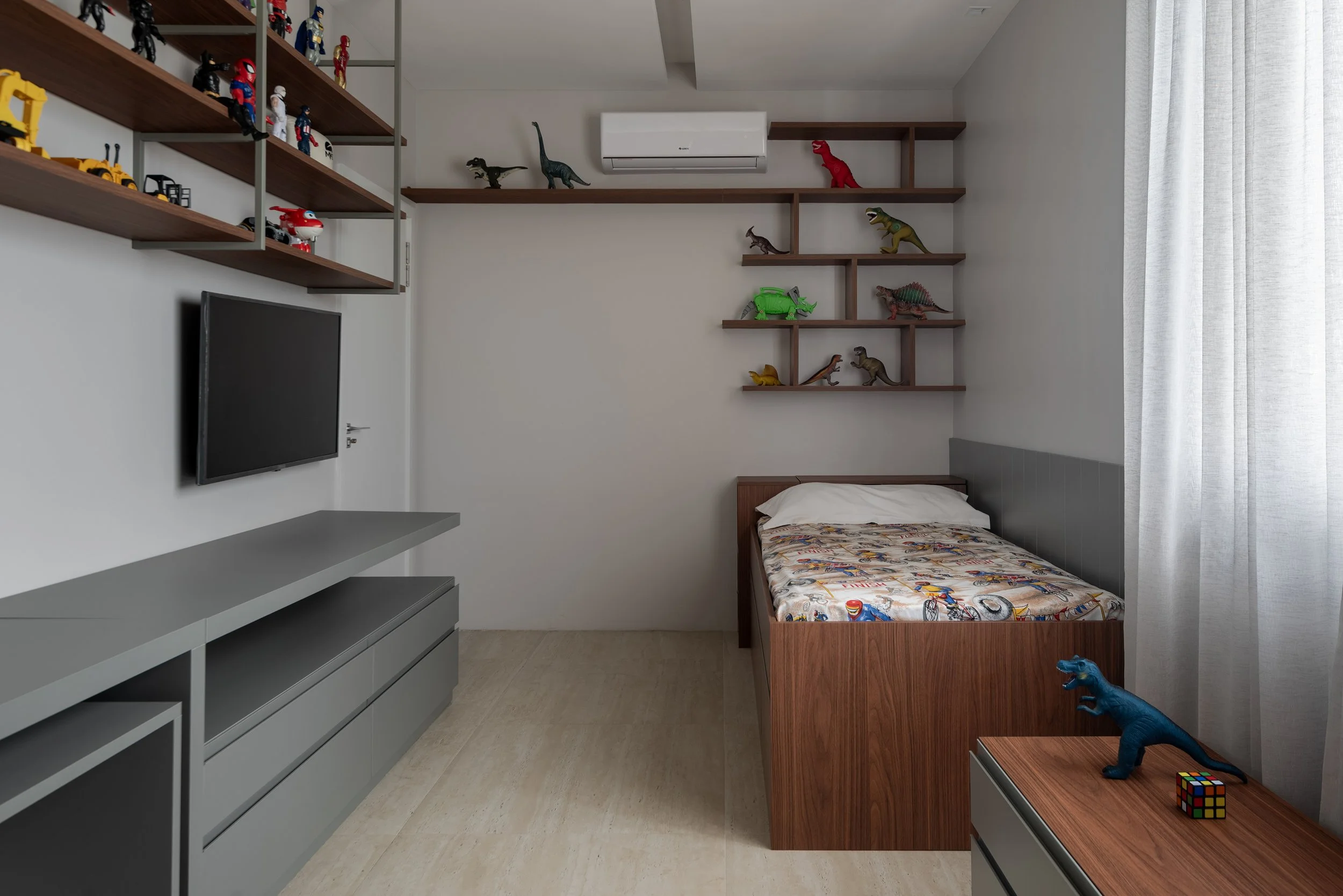 A bedroom with a bed, a flat-screen TV, a gray entertainment console, and shelves with Lego dinosaur and vehicle toys.
