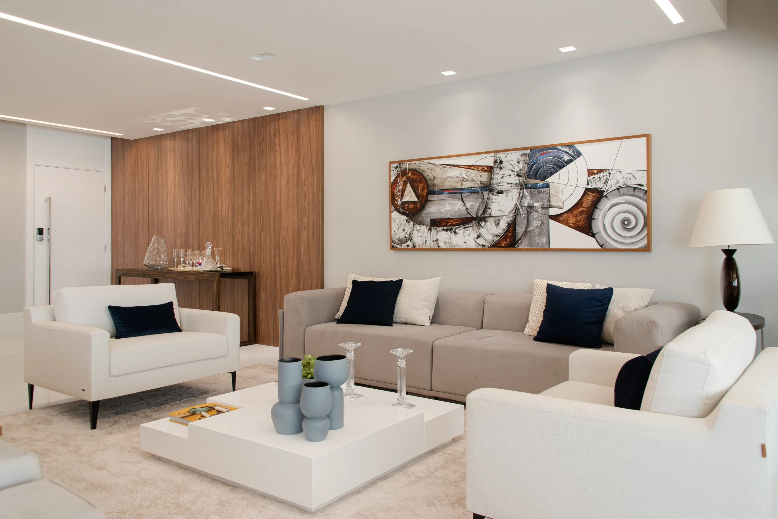 Modern living room with white, beige, and navy decor, abstract artwork, coffee table with vases and glasses, and a wooden accent wall.