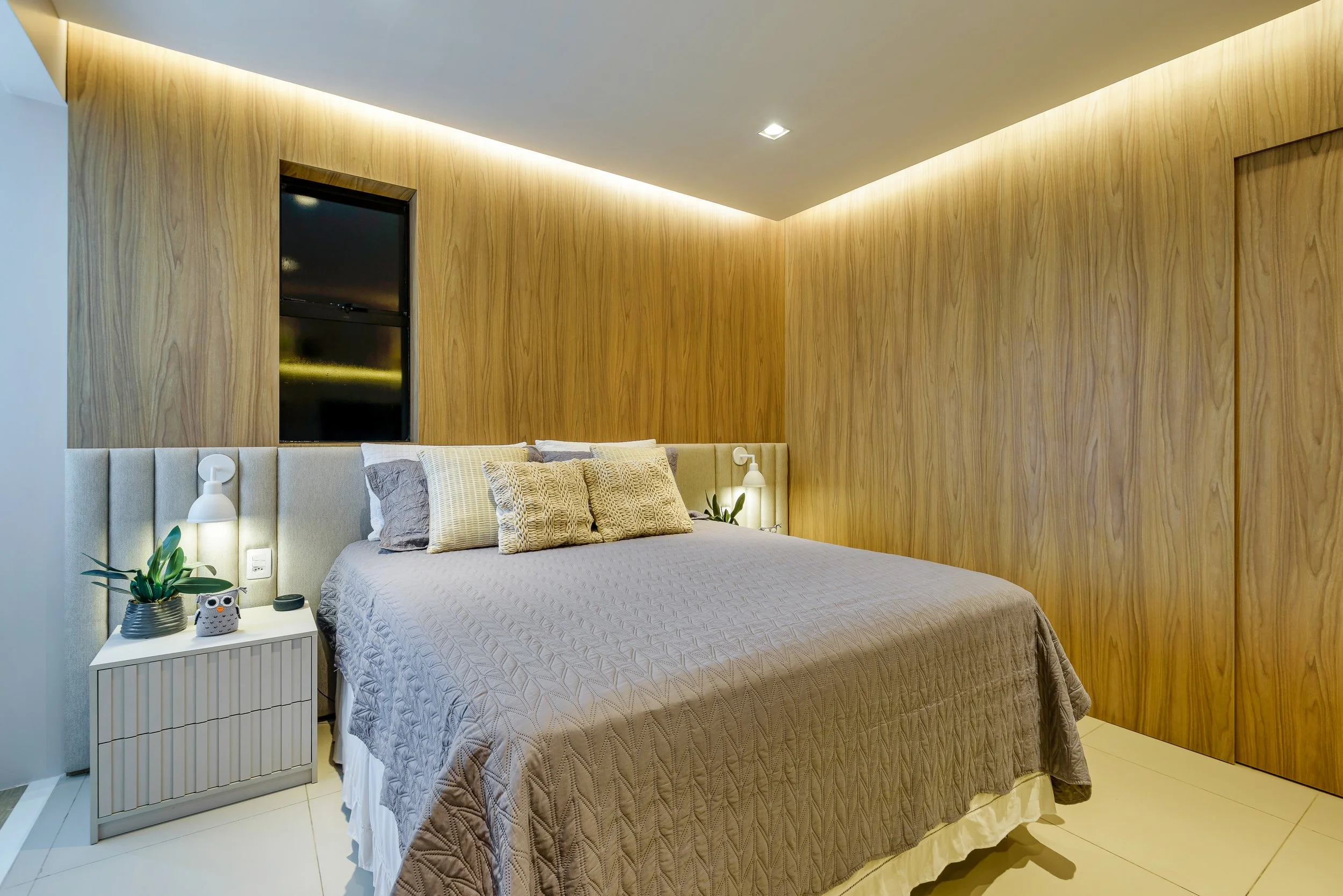 Modern bedroom with beige upholstered headboard, wooden paneled wall, and a bed with textured beige bedding and multiple pillows. White nightstand with a potted plant, owl figurine, and lamp, lit by soft lighting.