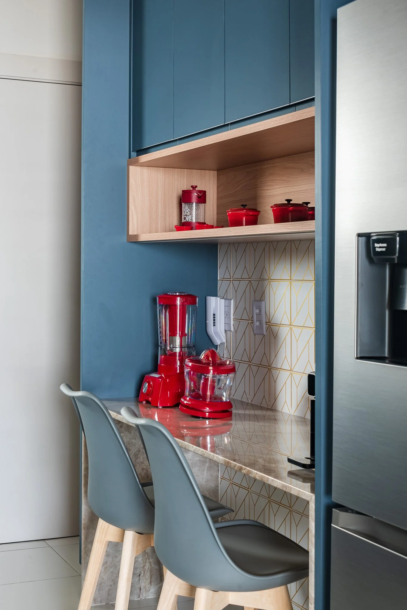 A modern kitchen corner with a blue cabinet and a beige marble countertop featuring a red blender and food processor. There are two gray chairs with wooden legs in front of the counter and yellow and white geometric patterned tiles on the wall behind