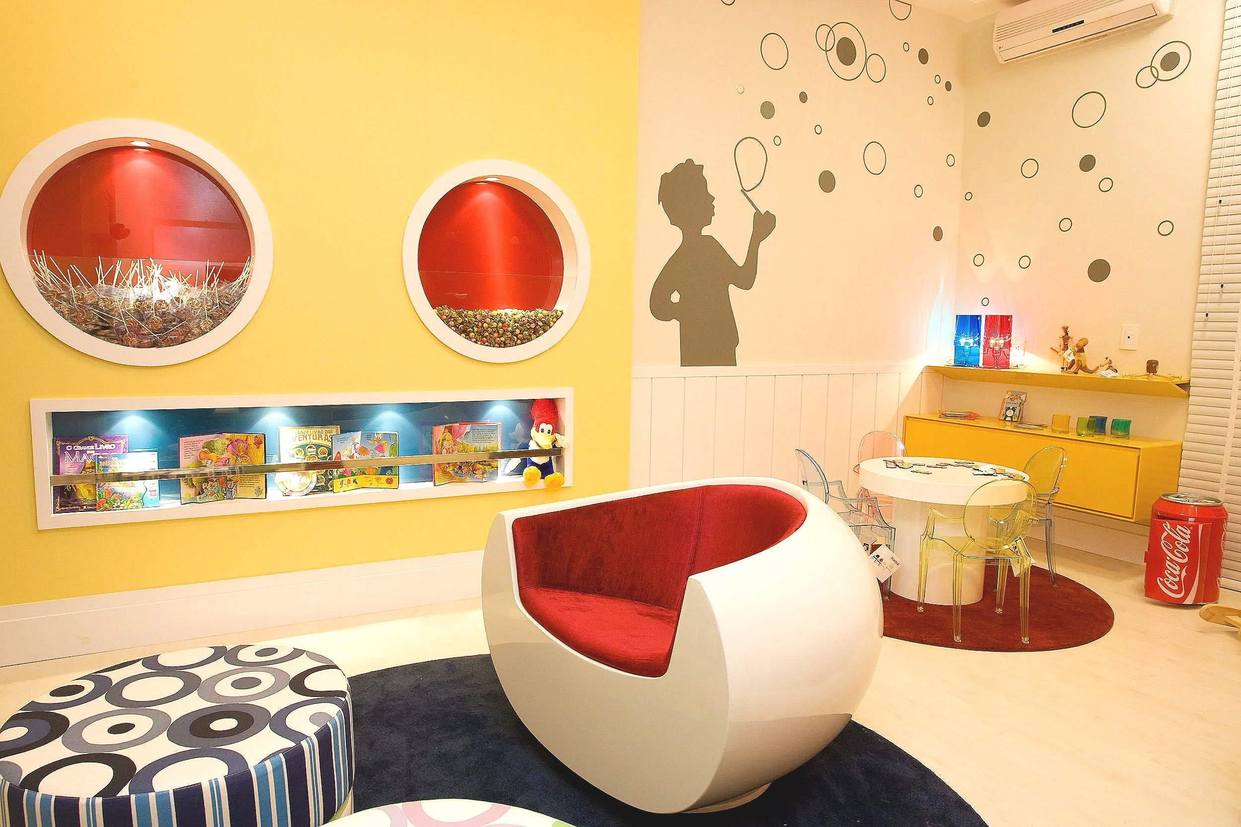 Colorful children's playroom with a yellow wall featuring round window designs and a silhouette mural of a girl playing with bubbles or a bubble wand, decorated with circle patterns. The room has modern furniture including a red and white chair, a sm