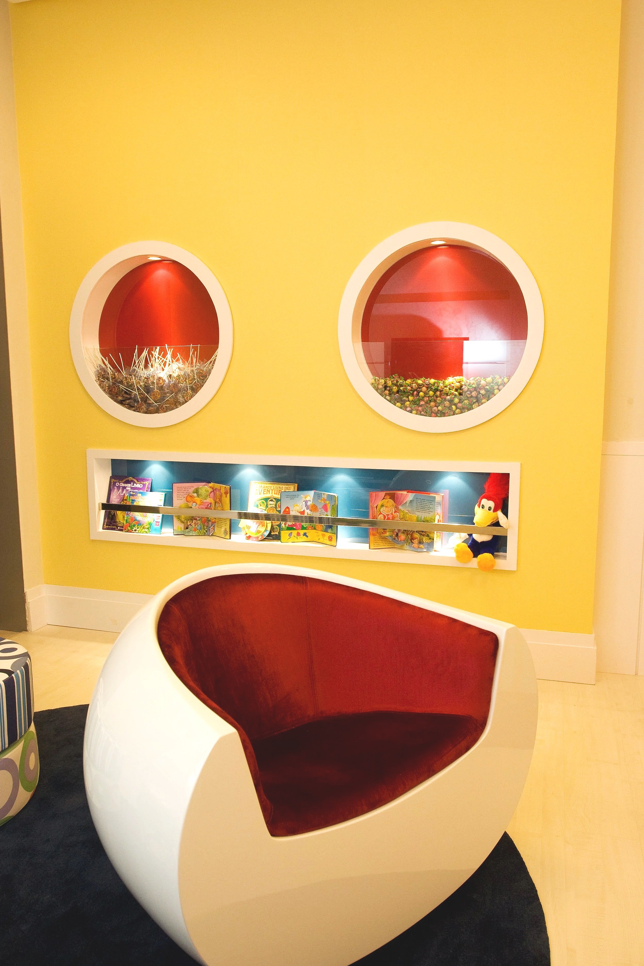 A modern, circular lounge chair with a brown interior in front of a yellow wall with two round display windows and a shelf with children's books and a plush toy.
