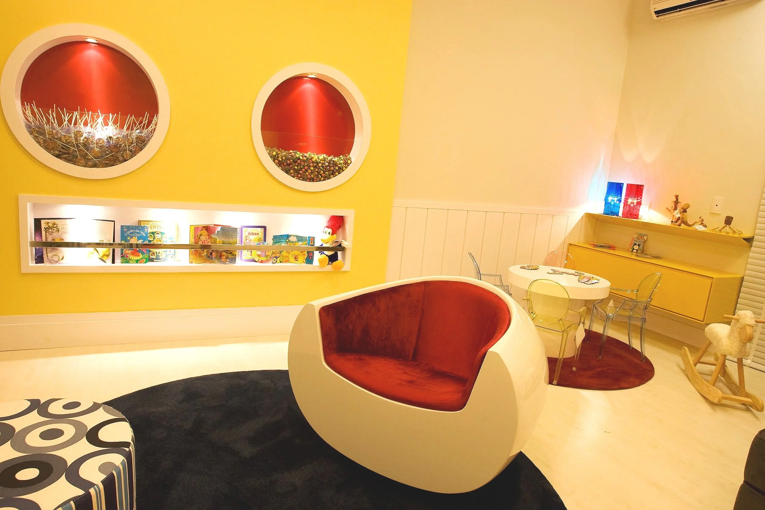Colorful living room with yellow walls, a red and white modern chair, a small round table with clear chairs, a yellow cabinet, and decorative elements including toys, books, and lights.