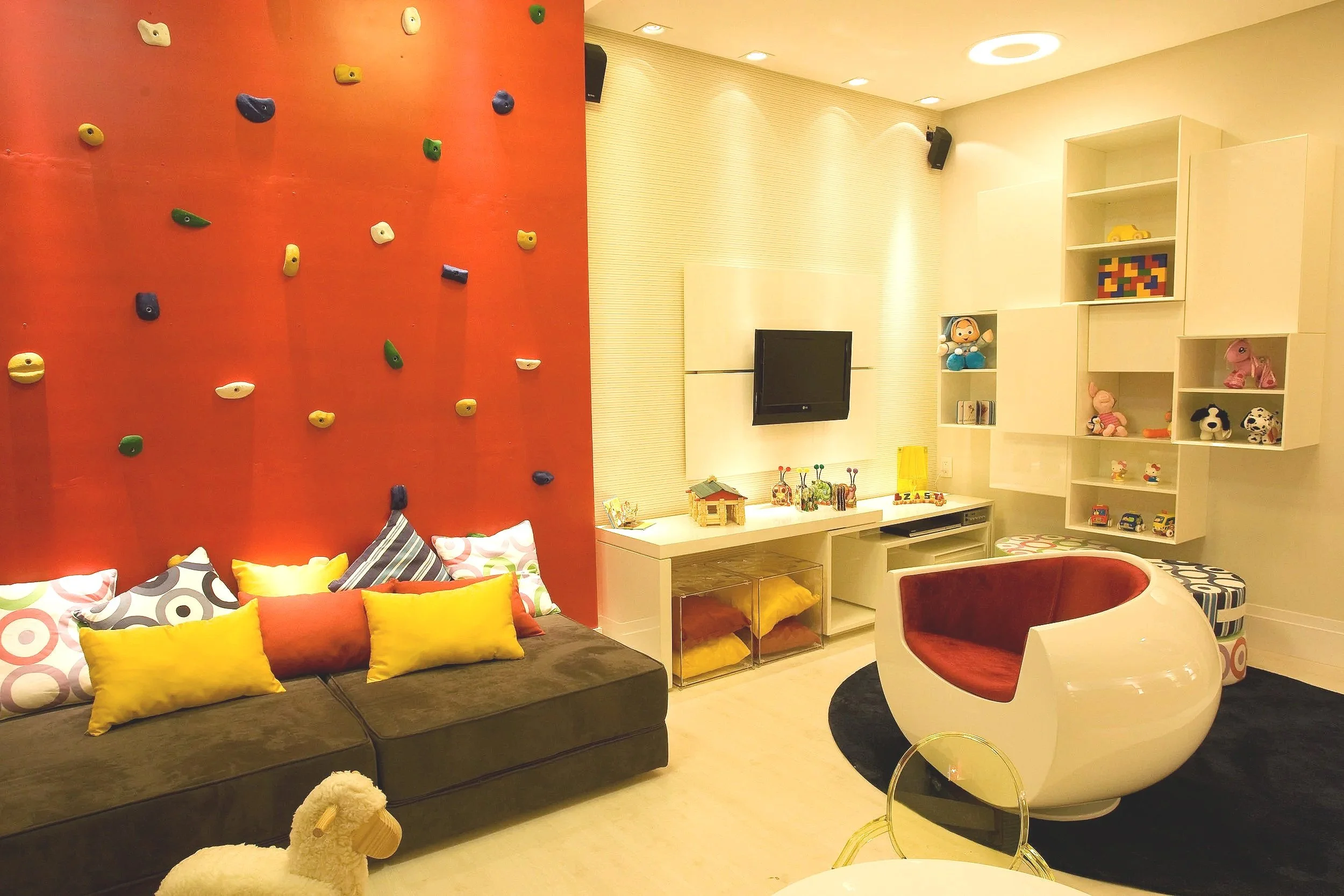 A colorful living room with a red climbing wall, a black sofa with multicolored pillows, a white round chair with red interior, and white shelving filled with toys and decorations, next to a wall-mounted TV.