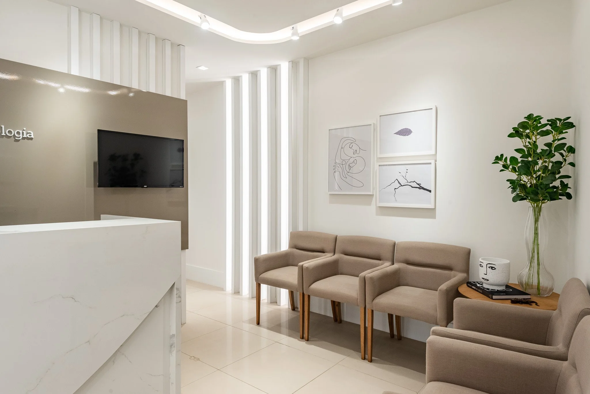 Minimalist waiting room with beige chairs, a potted plant, white walls, framed abstract art, and a flat-screen TV.