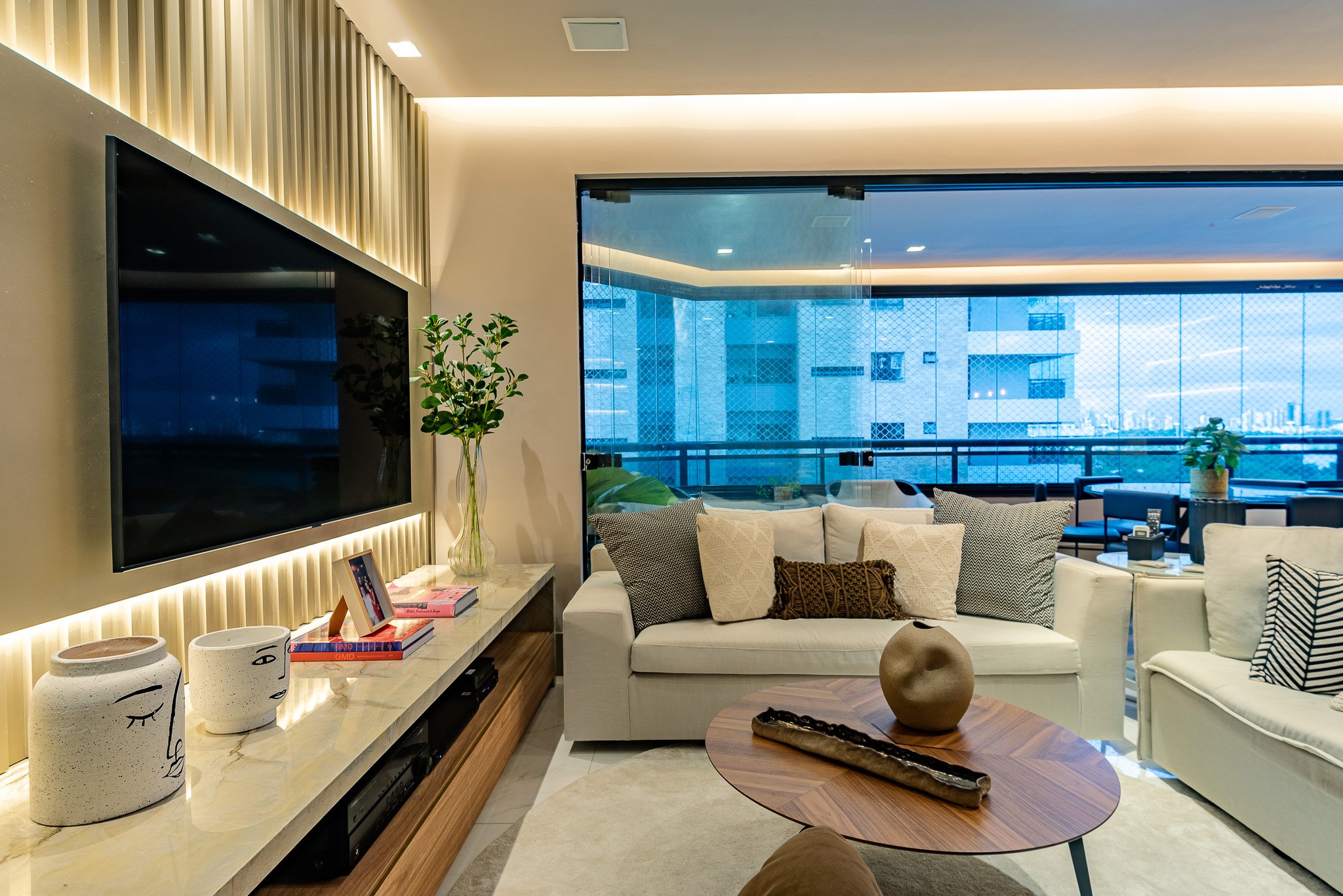 Modern living room with white sofas, decorative pillows, a wooden coffee table with a spherical sculpture, and a large flat-screen TV on the wall. Balcony with glass railing and city view in the background.