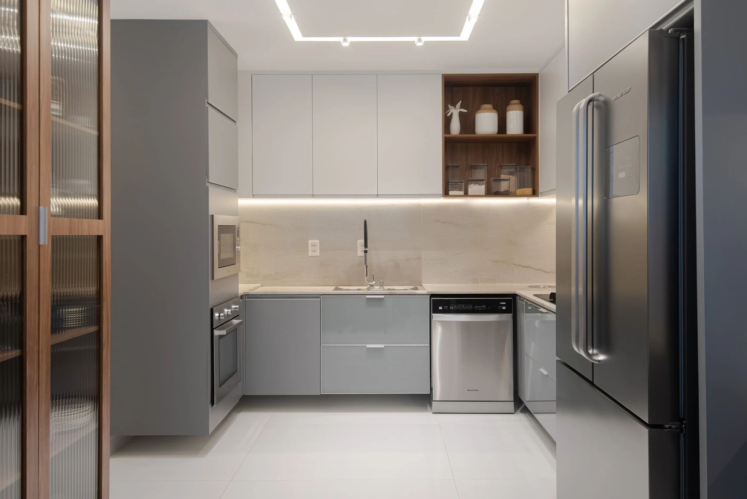 Modern kitchen with light gray cabinets, a white marble backsplash, and a dark wood open shelf with vases and jars. Stainless steel appliances include a refrigerator, oven, microwave, and dishwasher. The kitchen is well-lit with ceiling lighting and 