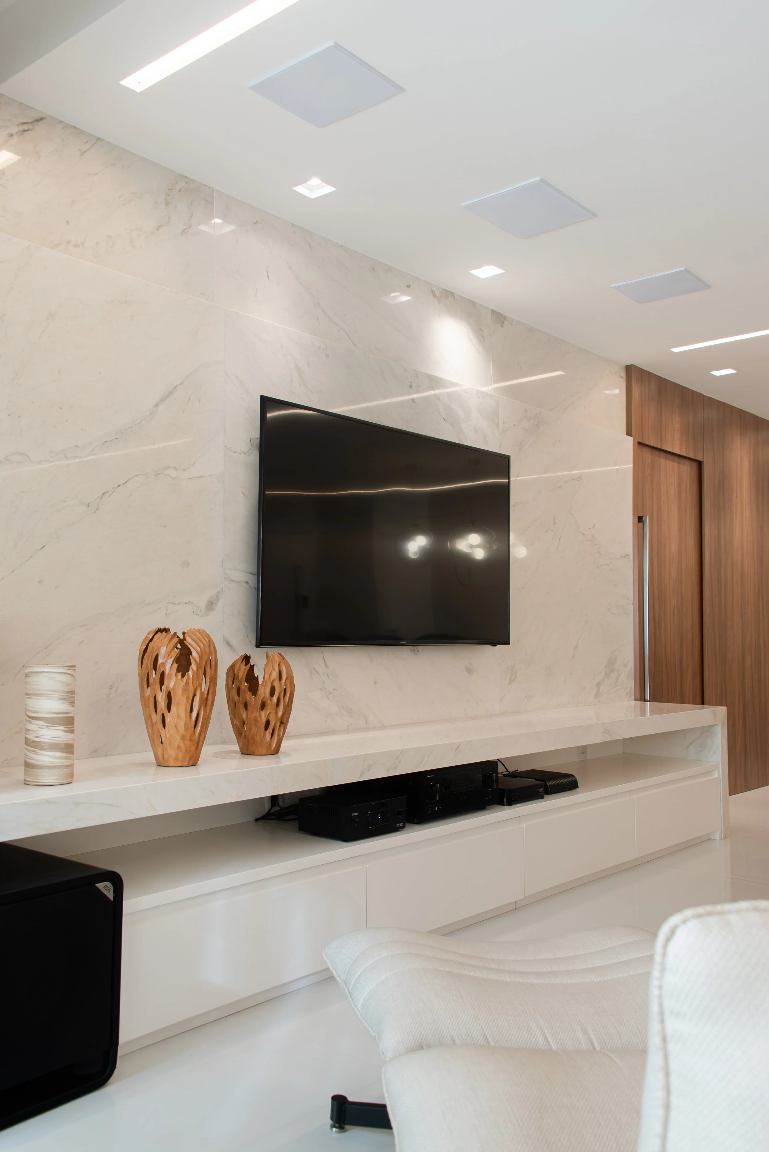 Modern living room with a mounted flat-screen TV on a marble wall, wooden decorative vases and a ceramic vase on a white media console, and part of a cream-colored chair visible in the foreground.
