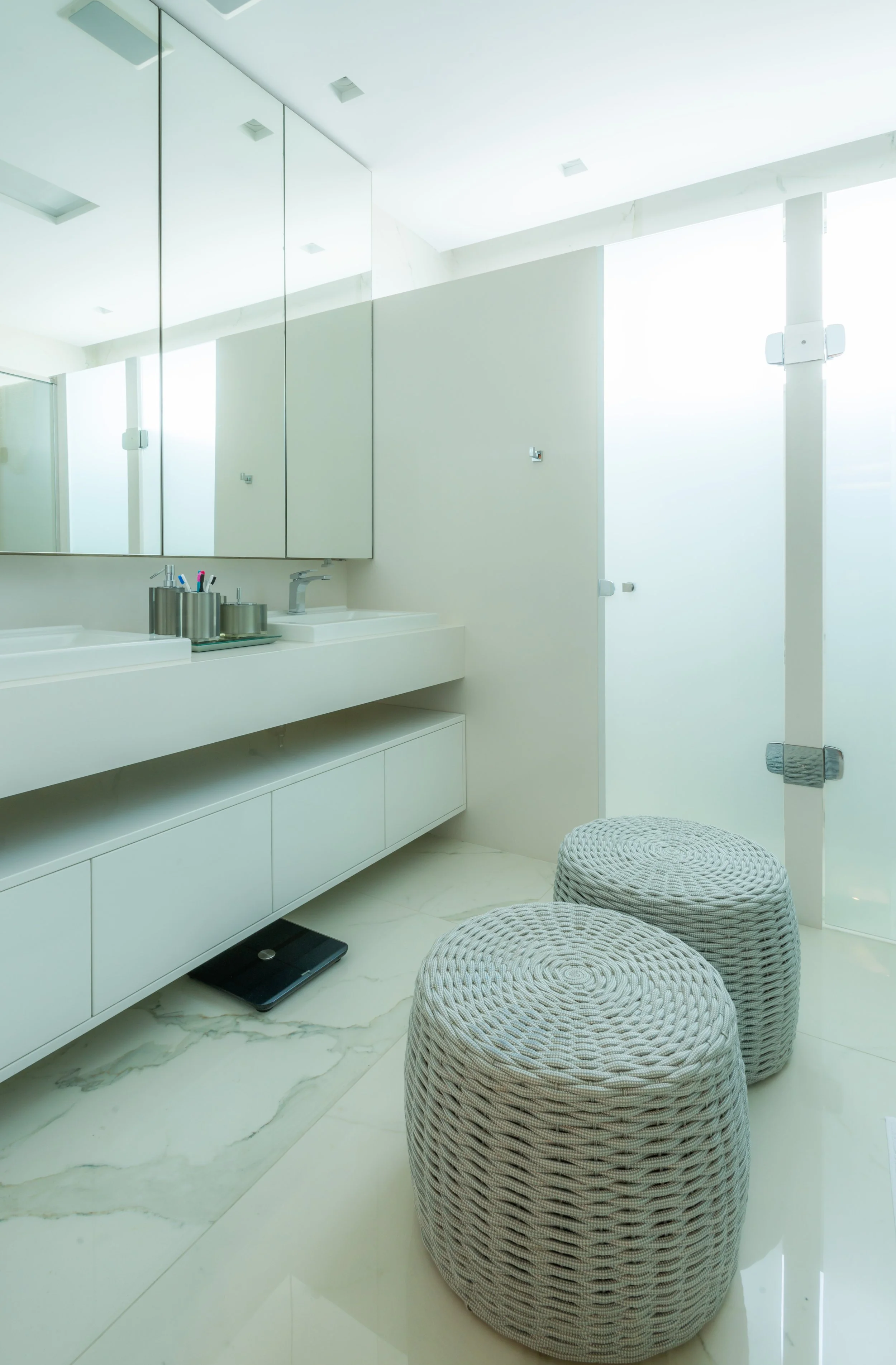 A modern, minimalistic bathroom with white cabinetry, a large mirror, light-colored marble flooring, two woven ottomans, and a frosted glass door letting in bright natural light.