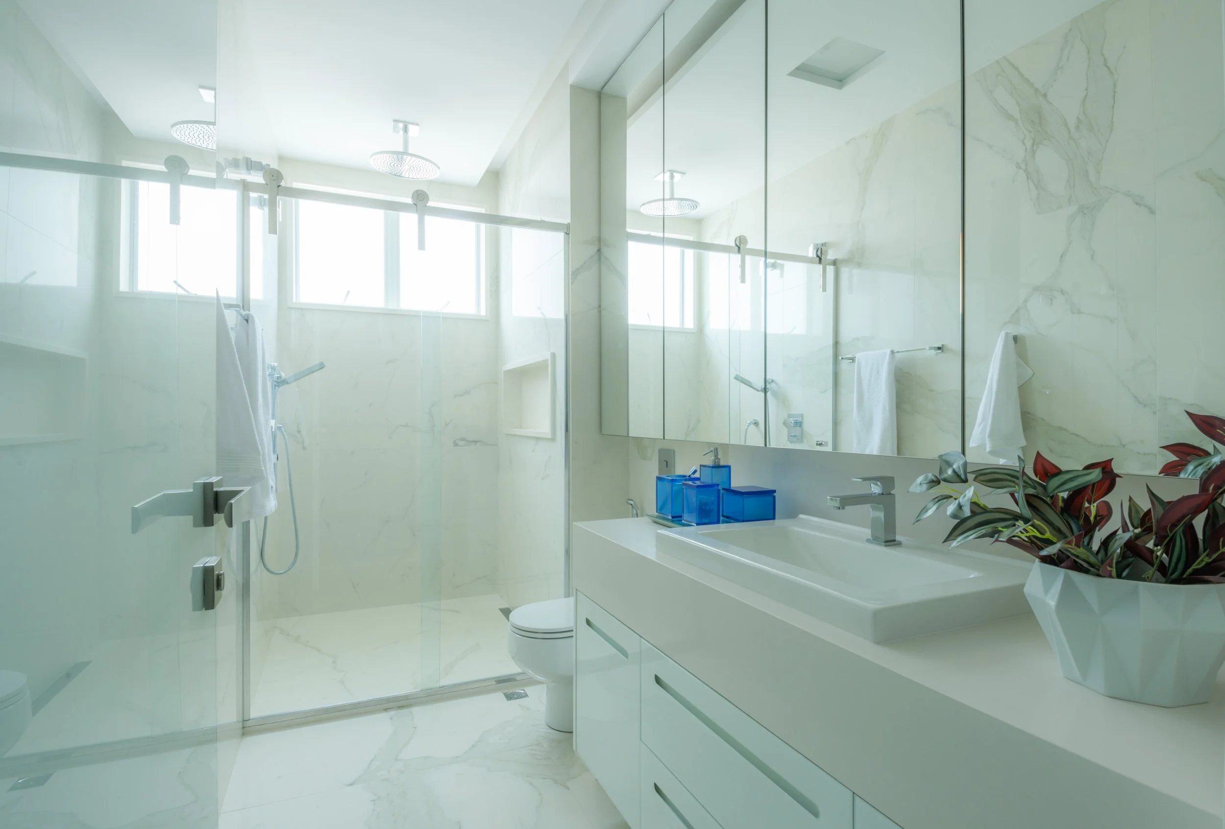 Modern bathroom with large mirror, white vanity, and walk-in shower with glass door, white marble walls, and multiple window panels for natural light.