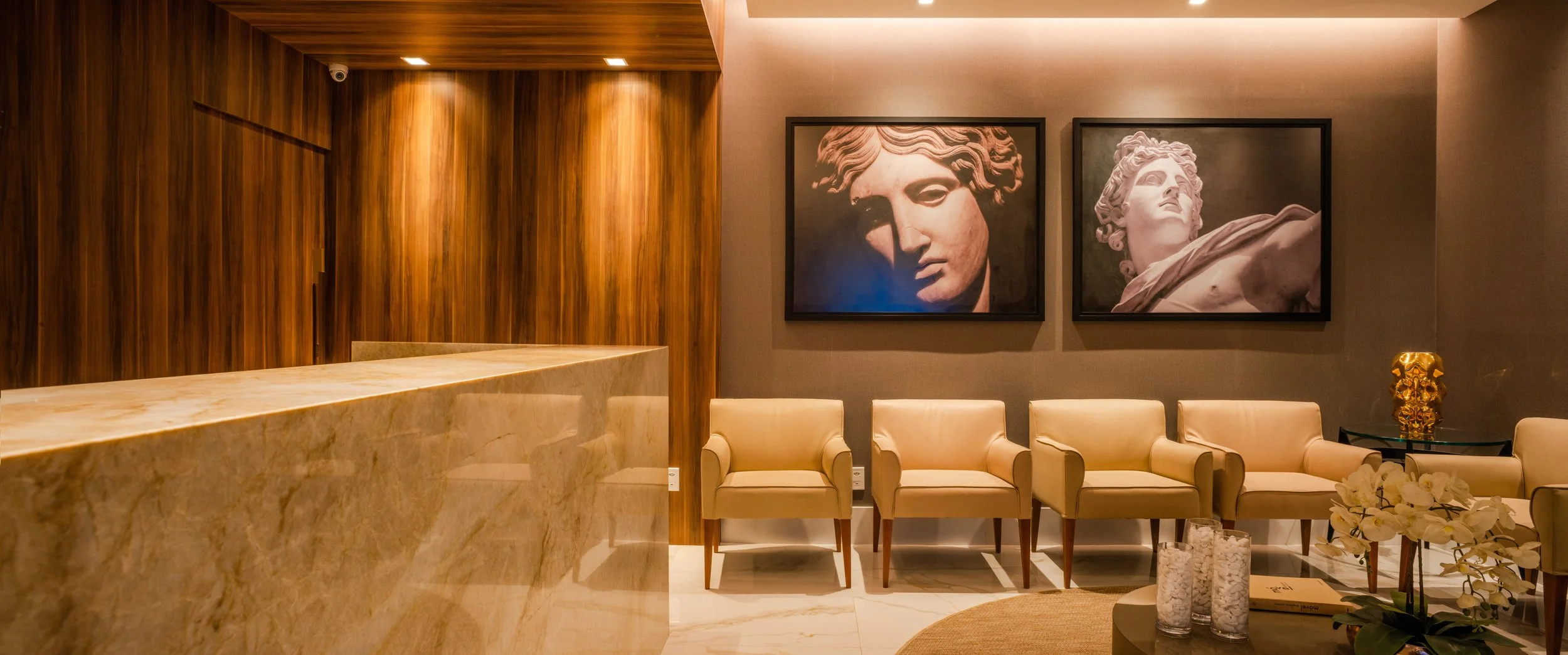 Modern hotel lobby with beige leather chairs, a marble reception desk, wall art featuring classical statues, and decorative items including a plant and candles.