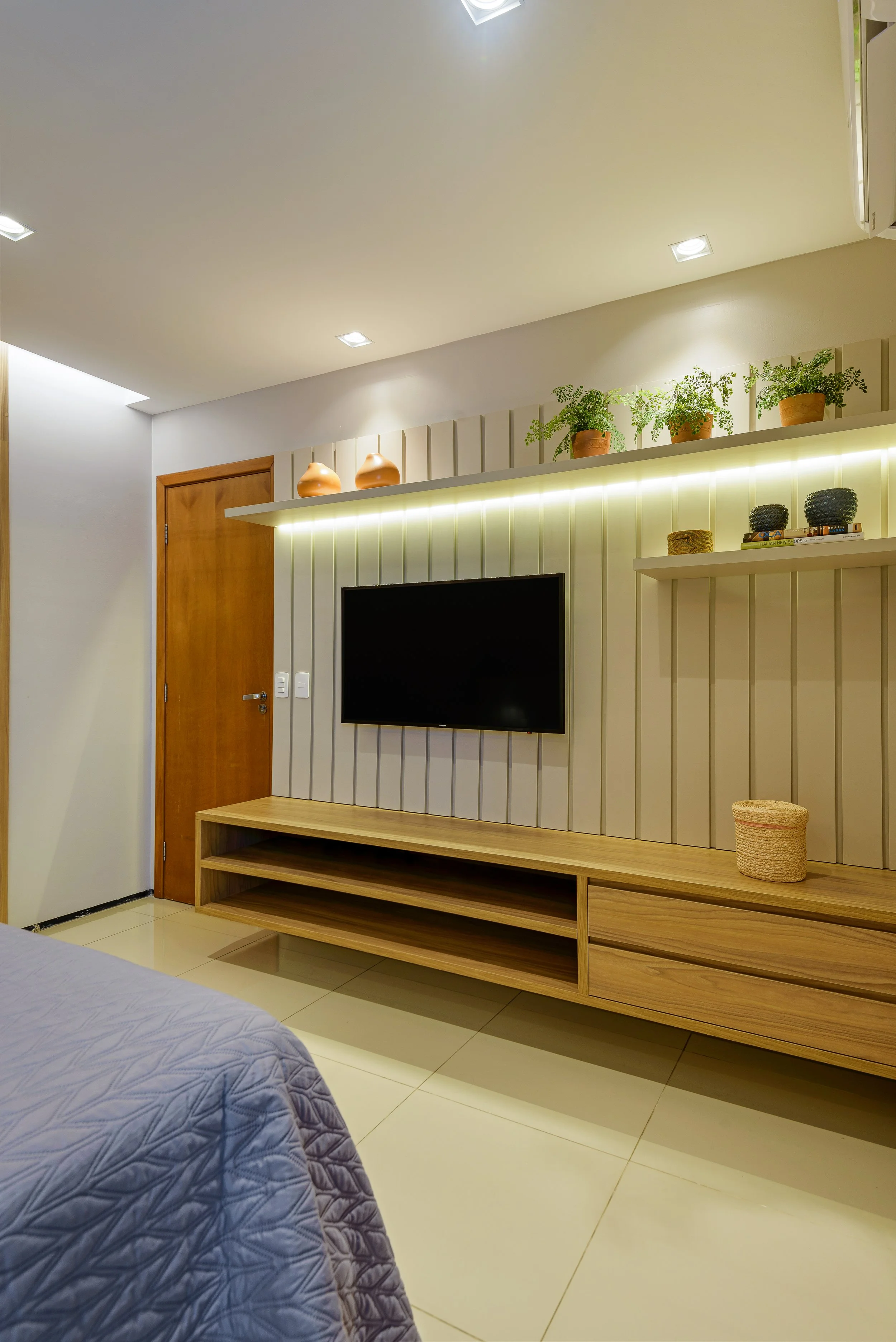 Modern bedroom with a flat-screen TV mounted on a neutral-colored wall, a wooden shelf below, and decorative plants and pottery on shelves above.