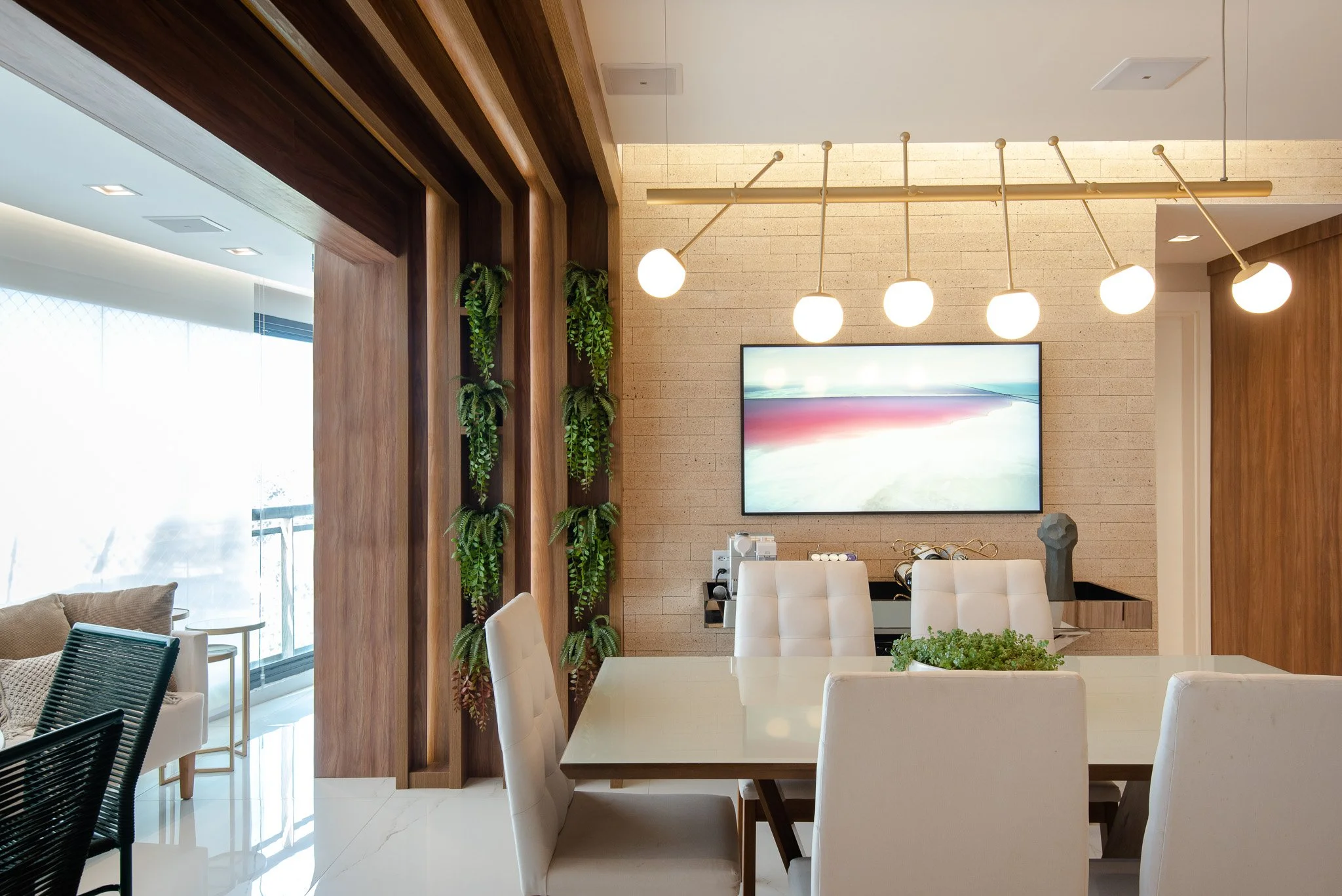 Modern dining room with a rectangular table and six white chairs, a wall-mounted flat-screen TV, wood paneling, hanging globe lights, and a vertical garden of green plants.