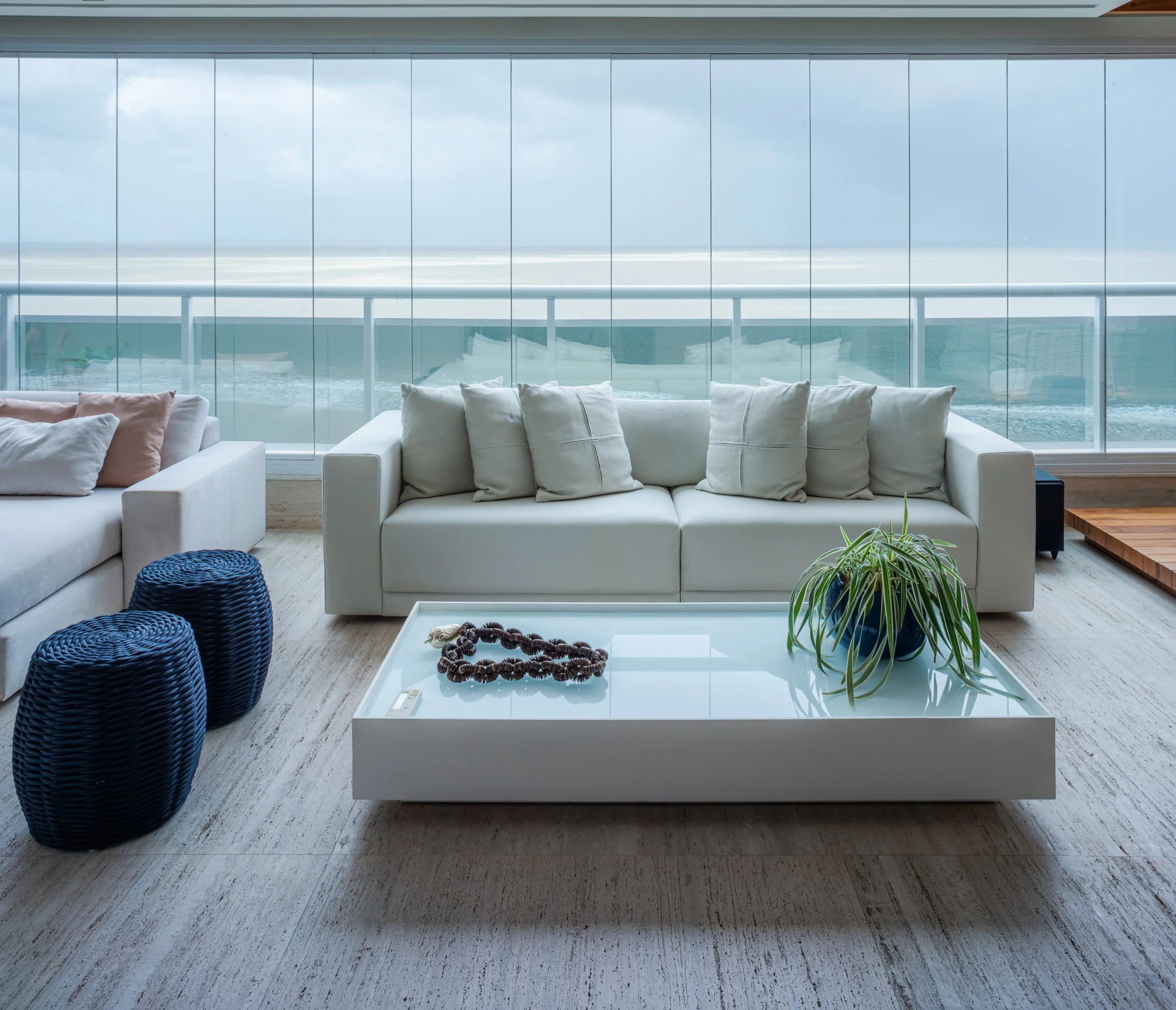 Modern living room with white sofa, decorative pillows, blue woven stools, glass coffee table with necklace and plant, and ocean view through large windows.