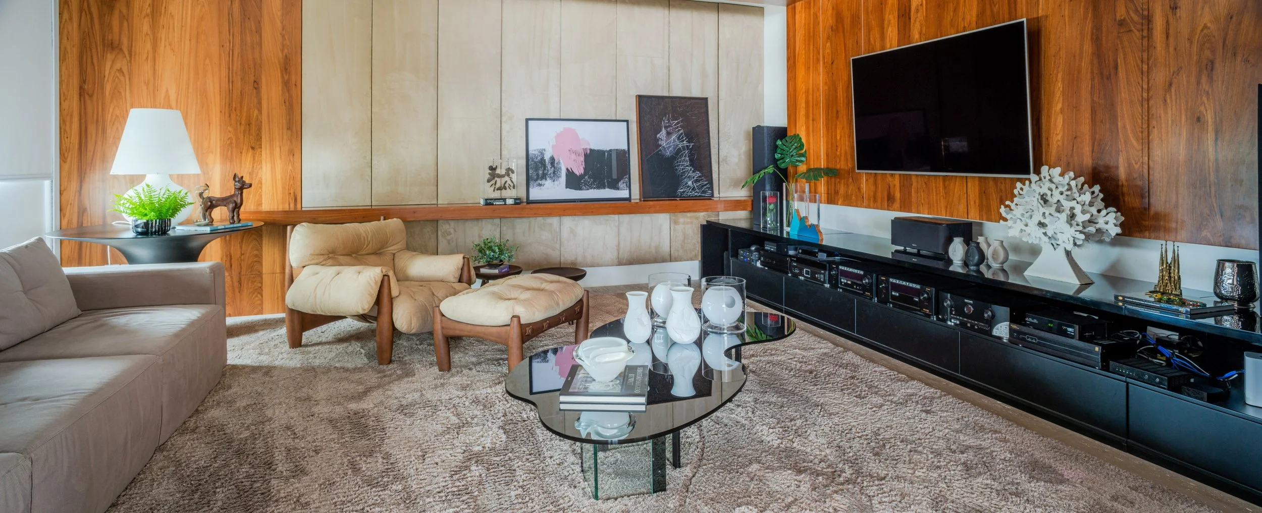 Living room with beige and brown furniture, a black entertainment center with electronic devices, a mounted large TV, a glass coffee table with decorative vases and books, and artwork on the wood-paneled walls.