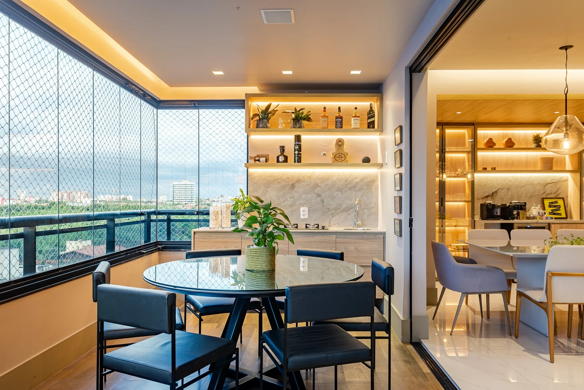 Modern apartment balcony with a glass table, black chairs, a potted plant, and a view of city buildings and greenery, separated from the indoor space by a sliding door.