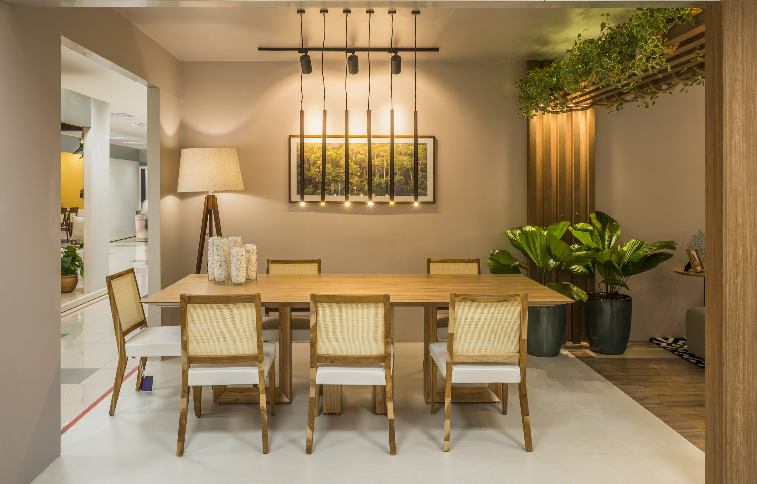 Modern dining room with a wooden table and six chairs, large potted plants, a floor lamp, and decorative candles, with artwork and lighting fixtures on the wall.
