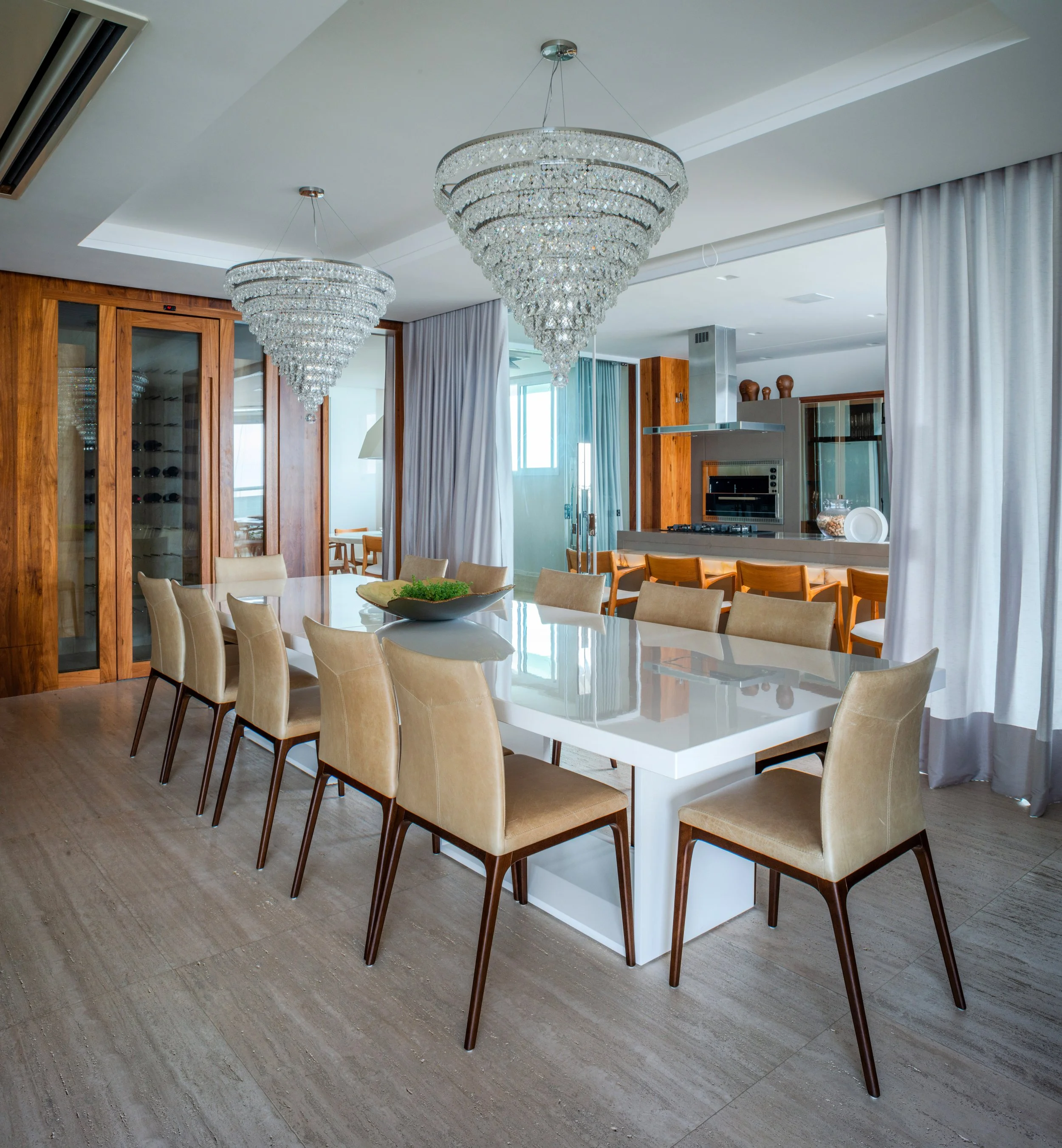 Modern dining room with a white table, beige chairs, crystal chandeliers, and a view of the open kitchen with wooden cabinets and seating area.
