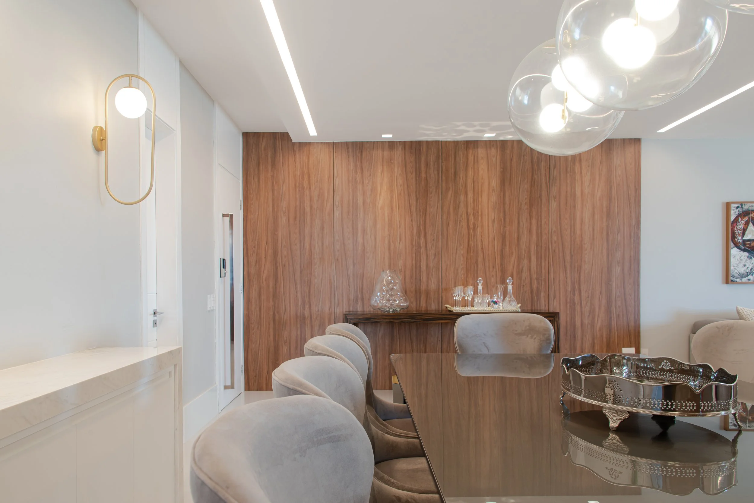 Modern dining room with a polished wooden table, beige upholstered chairs, a wooden accent wall, contemporary glass pendant lights, and a wooden sideboard with glass decanters and decorative items.