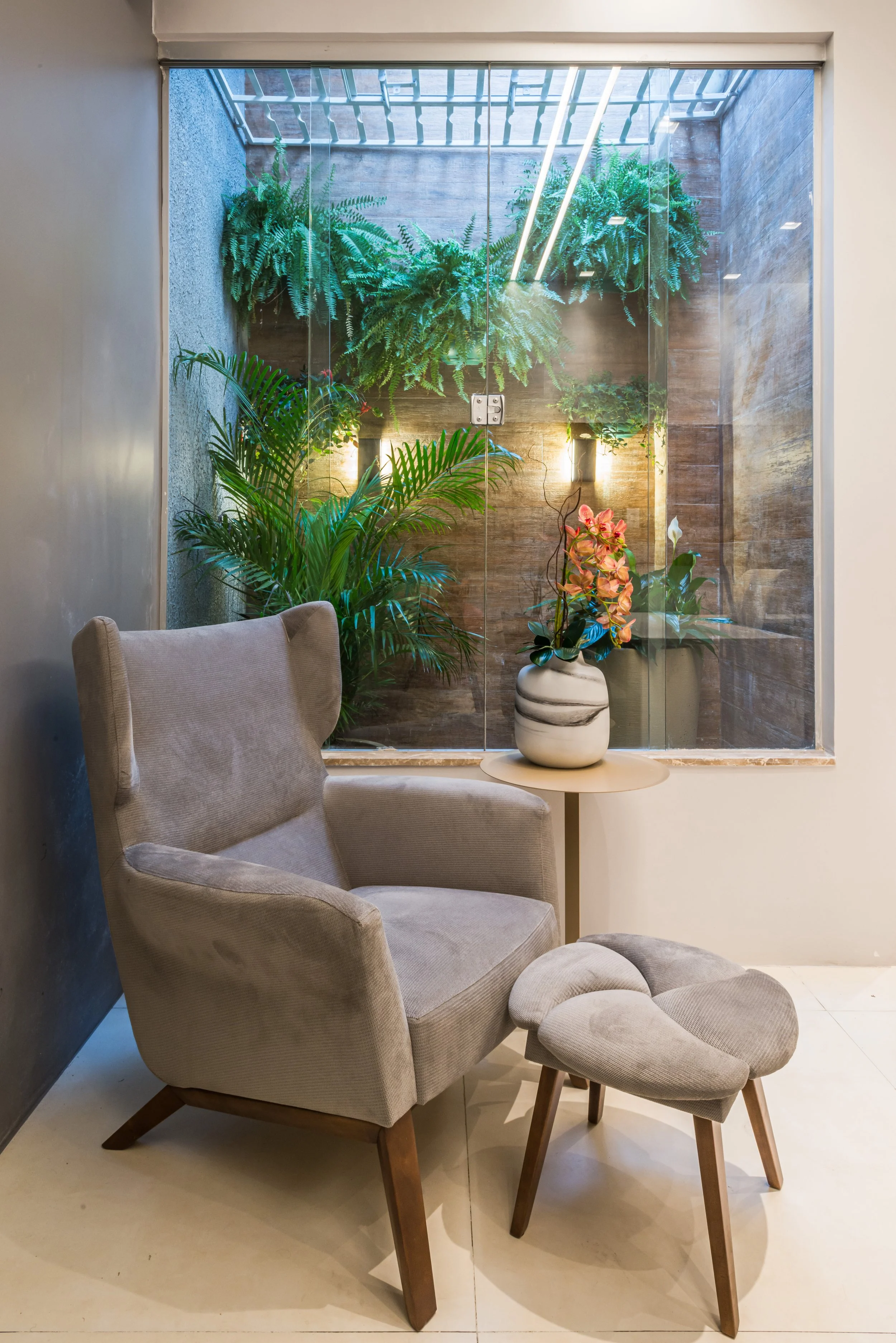 A cozy corner with a beige armchair and matching ottoman next to a small round side table holding a white and grey vase with pink and green flowers. Behind the chair, a large window reveals a small indoor garden with lush green plants and ferns, and 